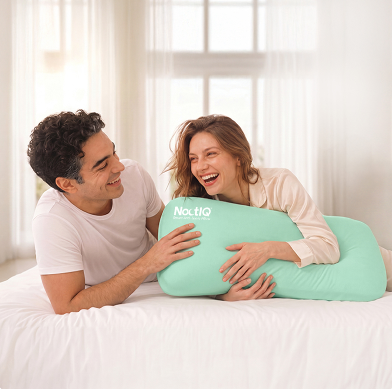 A laughing couple on a bed, holding a mint green NoctIQ Smart Anti-Snore Pillow.