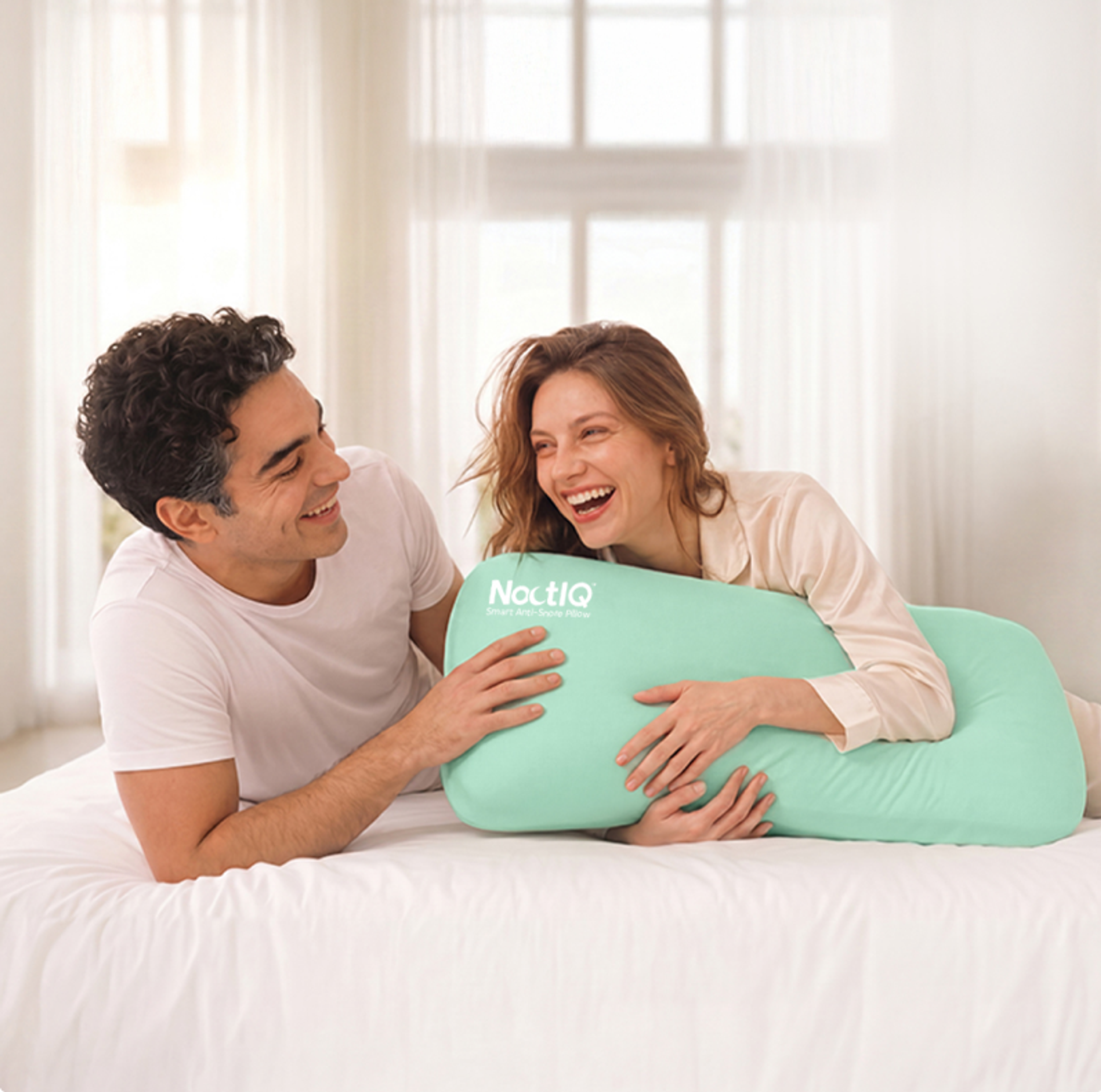 A laughing couple on a bed, holding a mint green NoctIQ Smart Anti-Snore Pillow.