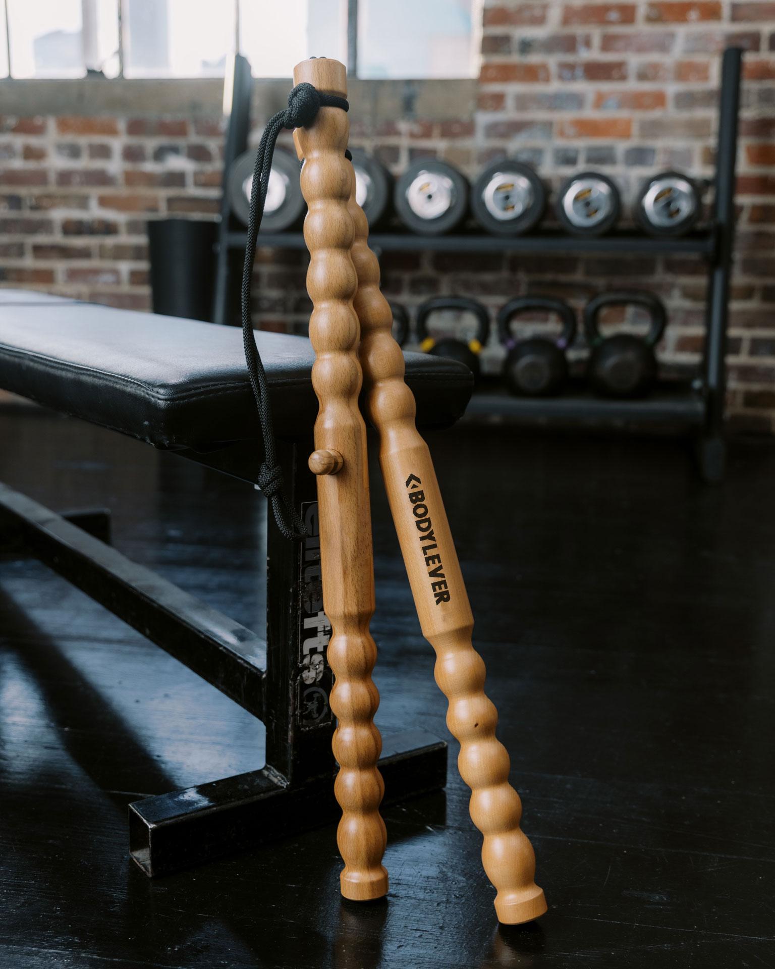 Two segmented wooden BODYLEVER fitness tools with a connecting cord, leaning against a gym bench.