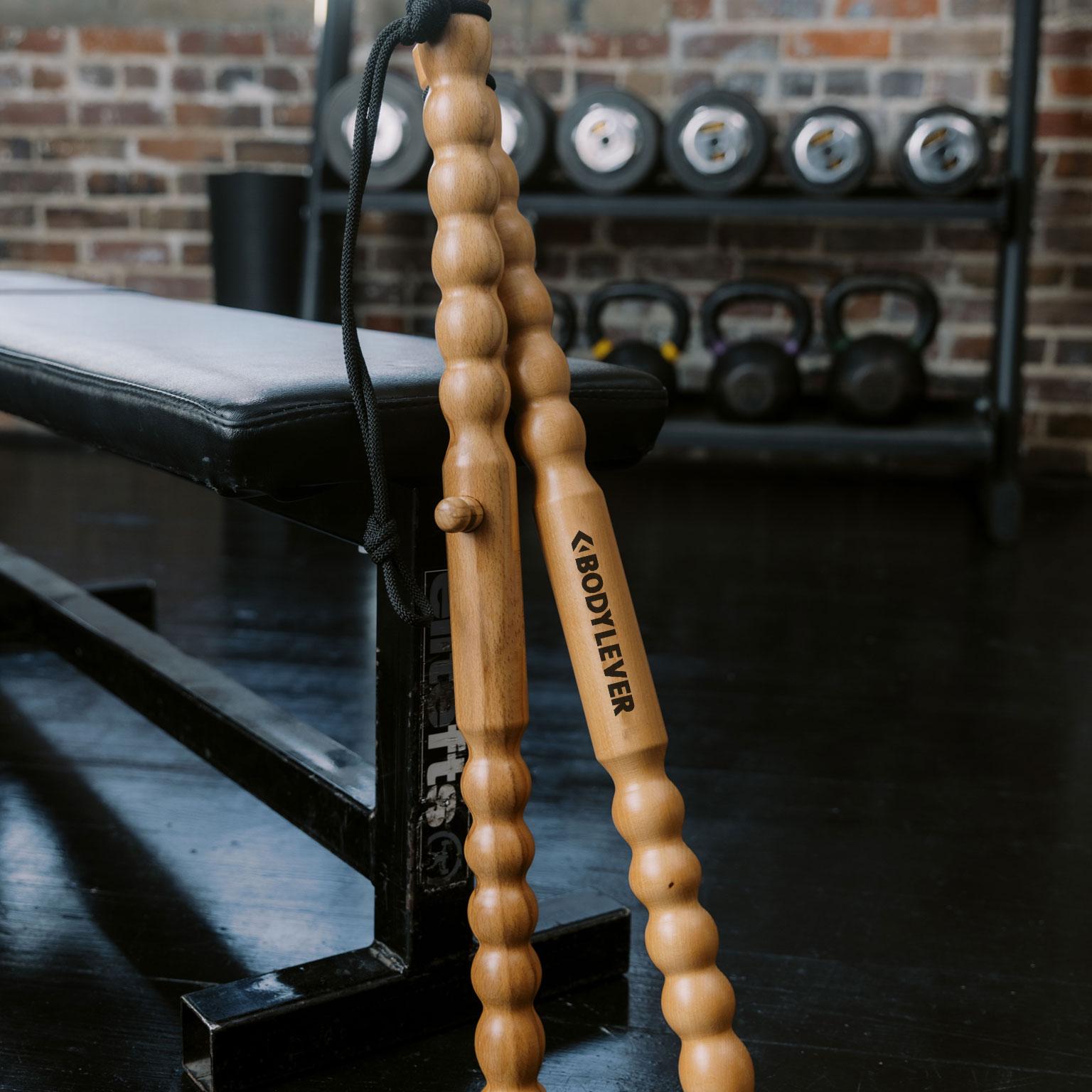 Two segmented wooden BODYLEVER fitness tools with a connecting cord, leaning against a gym bench.