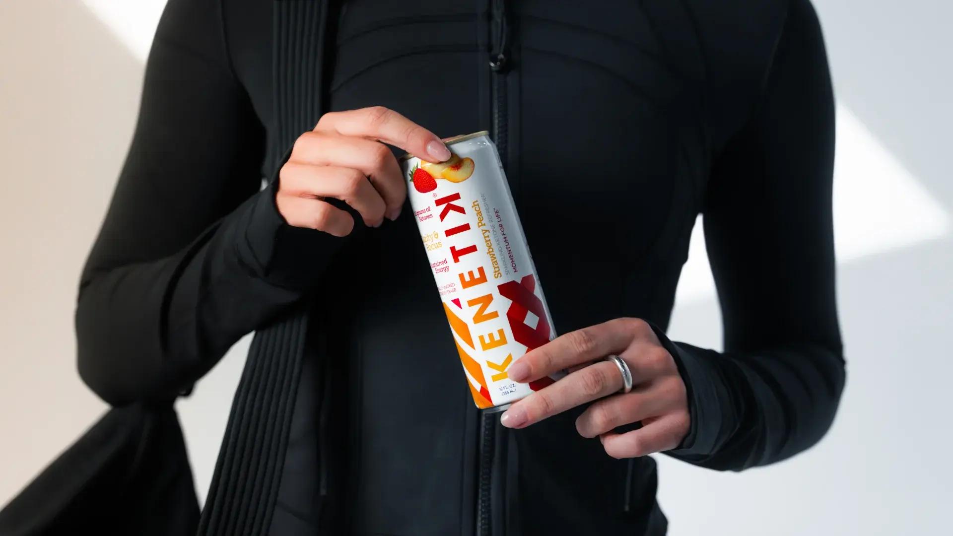 A person in black athletic wear holds a can of KENETIX Strawberry Peach drink.