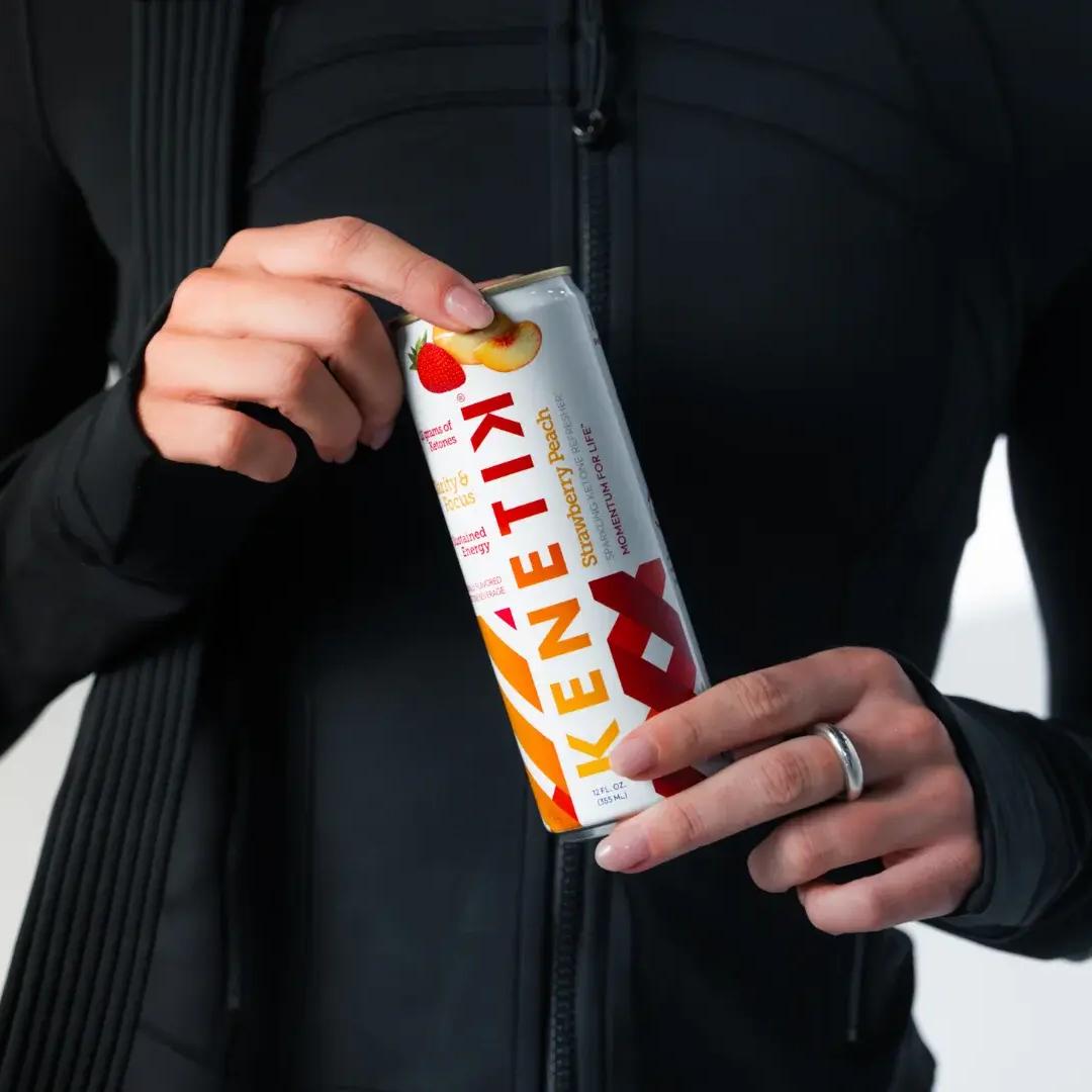 A person in black athletic wear holds a can of KENETIX Strawberry Peach drink.