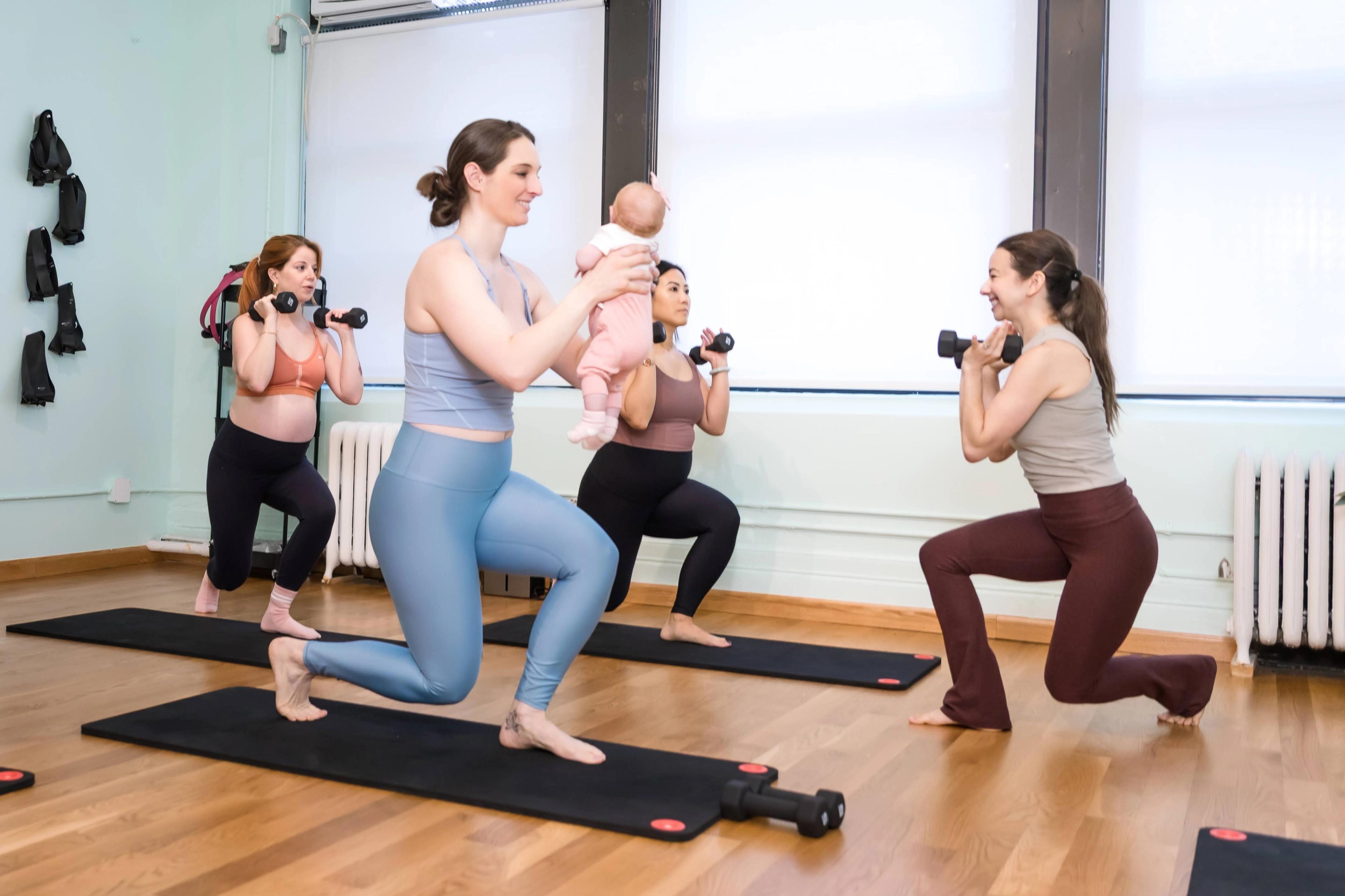 Four women, including one pregnant and one holding a baby, perform lunges with dumbbells in a fitness class.