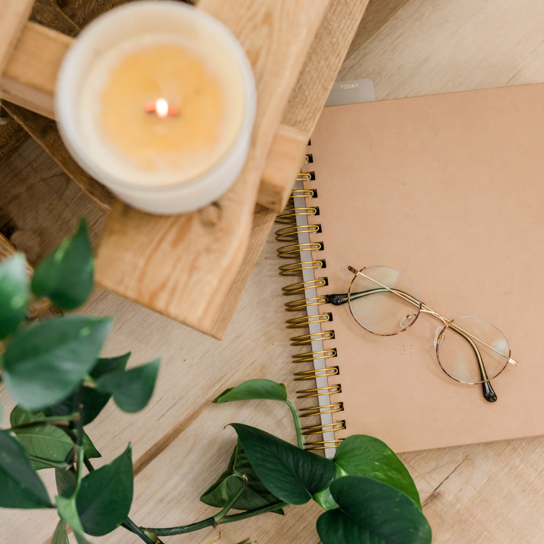 A lit candle on a wooden stand next to a brown spiral notebook with gold-rimmed glasses, and a green plant on a light wooden surface.