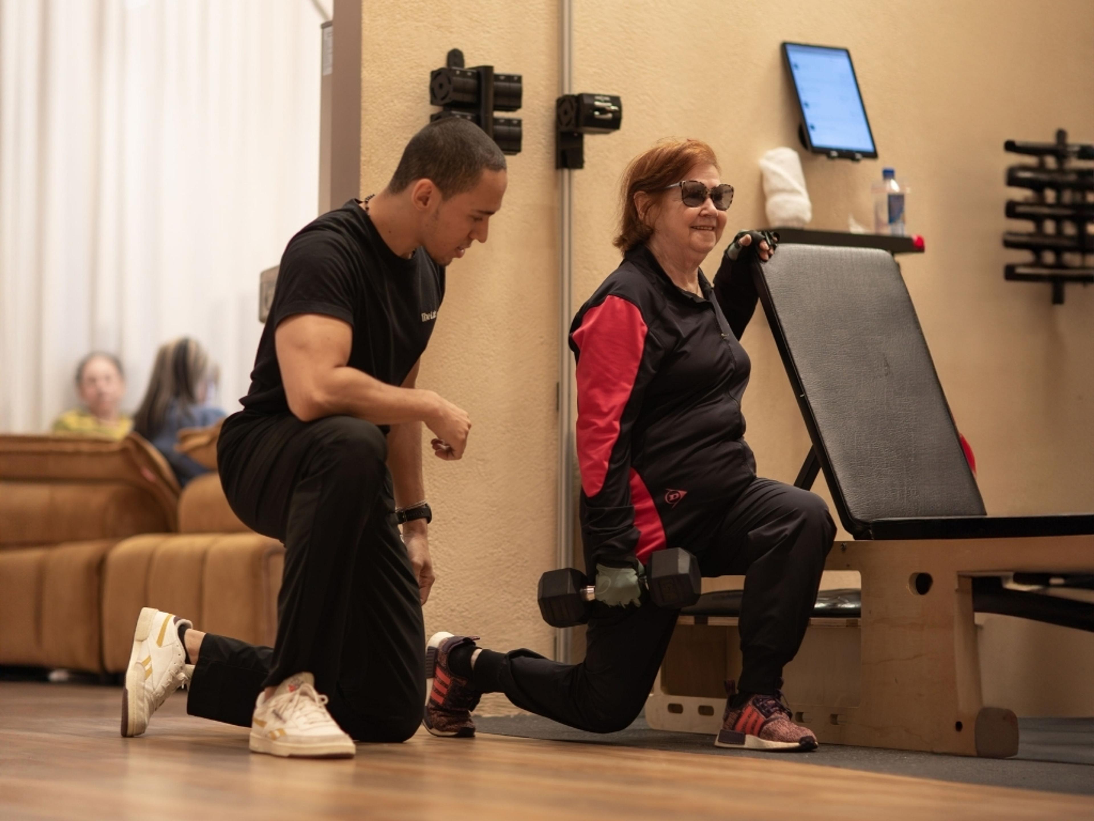 An older woman in sunglasses smiles while doing lunges with a dumbbell, supervised by a male personal trainer.