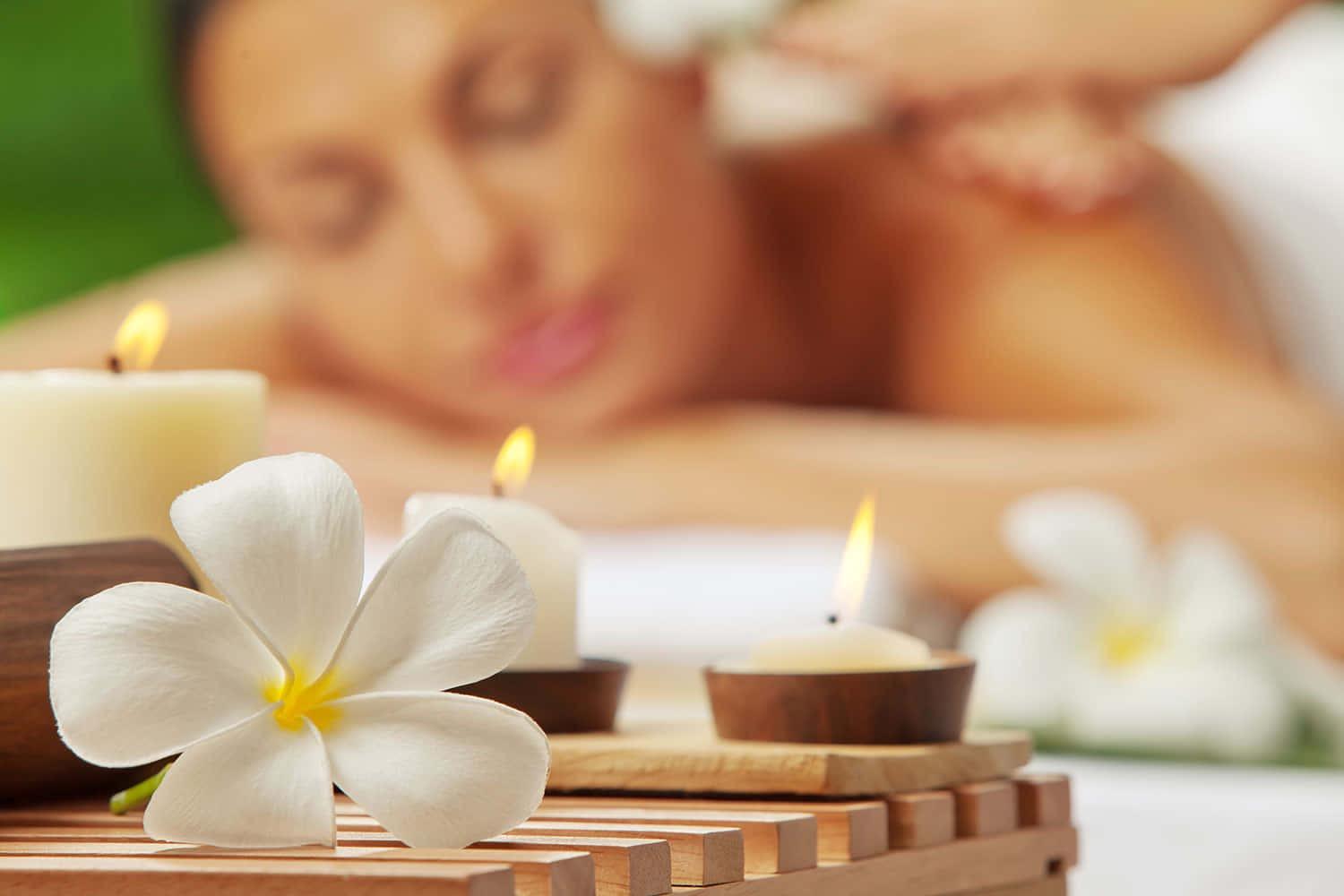 Close-up of a white plumeria flower and lit candles with a blurred background of a woman receiving a massage.
