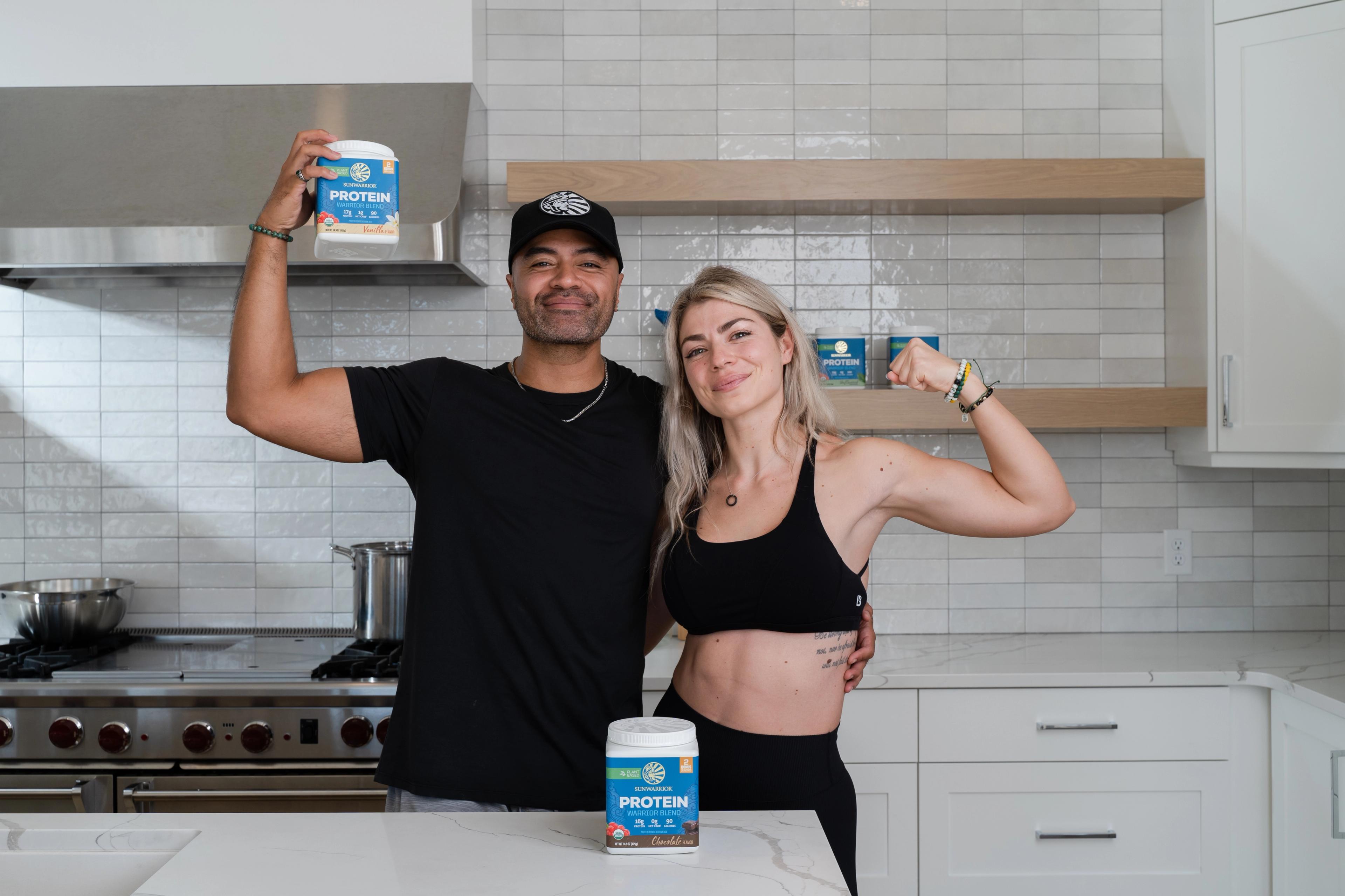 A man holds up a "PROTEIN BAR" product while a woman flexes her bicep in a modern kitchen.