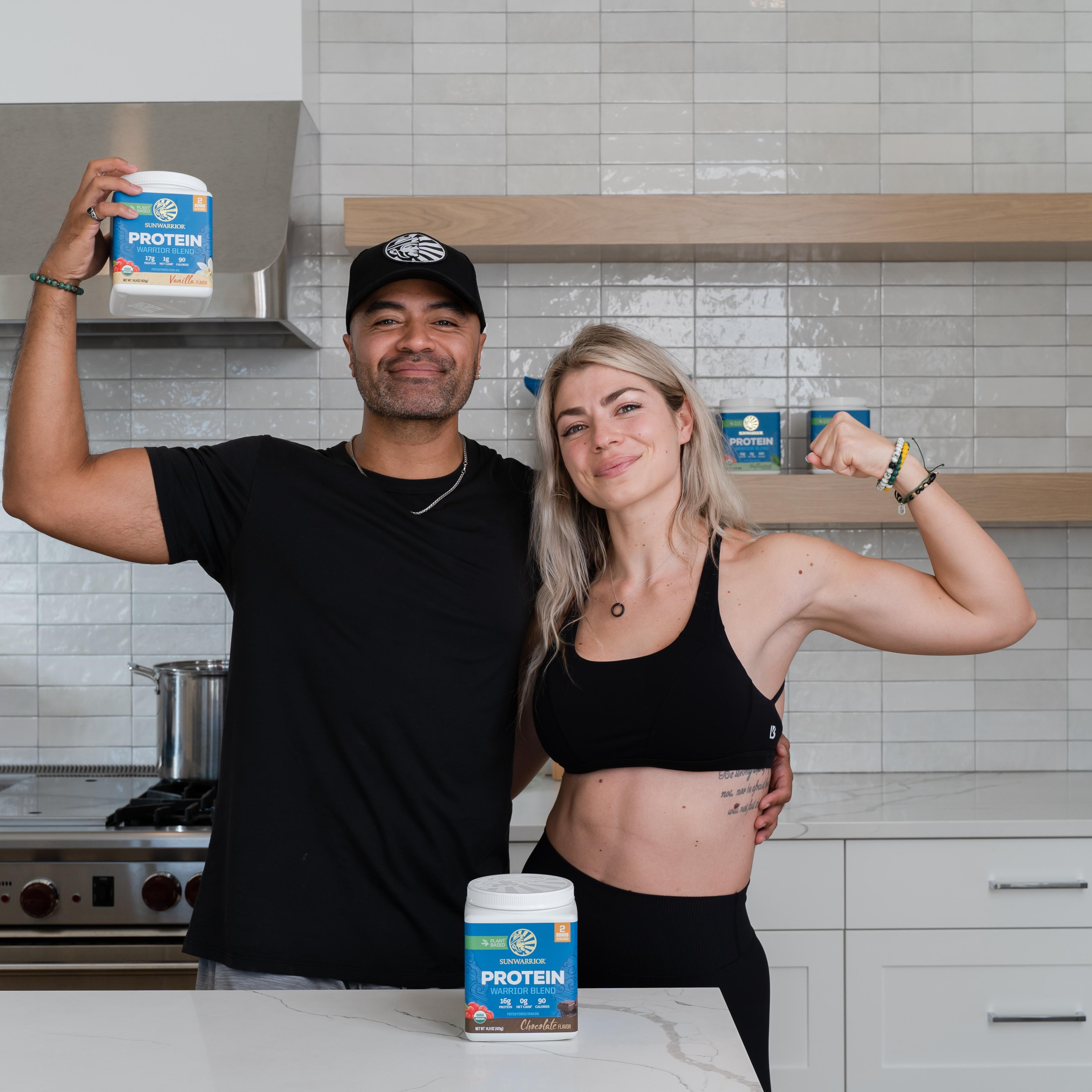 A man holds up a "PROTEIN BAR" product while a woman flexes her bicep in a modern kitchen.