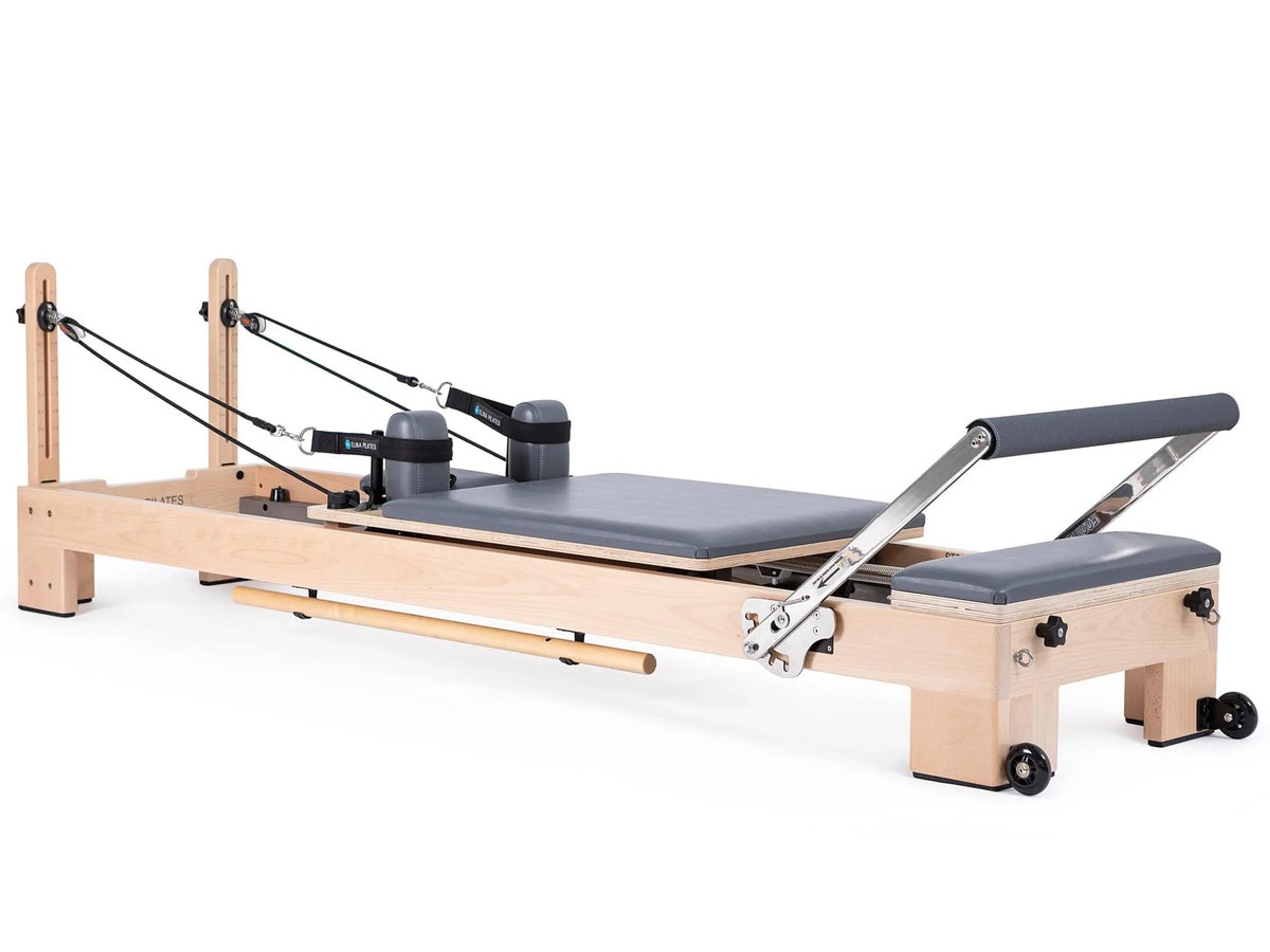 A Pilates reformer machine with a light wood frame and gray padding, featuring ropes and a foot bar.