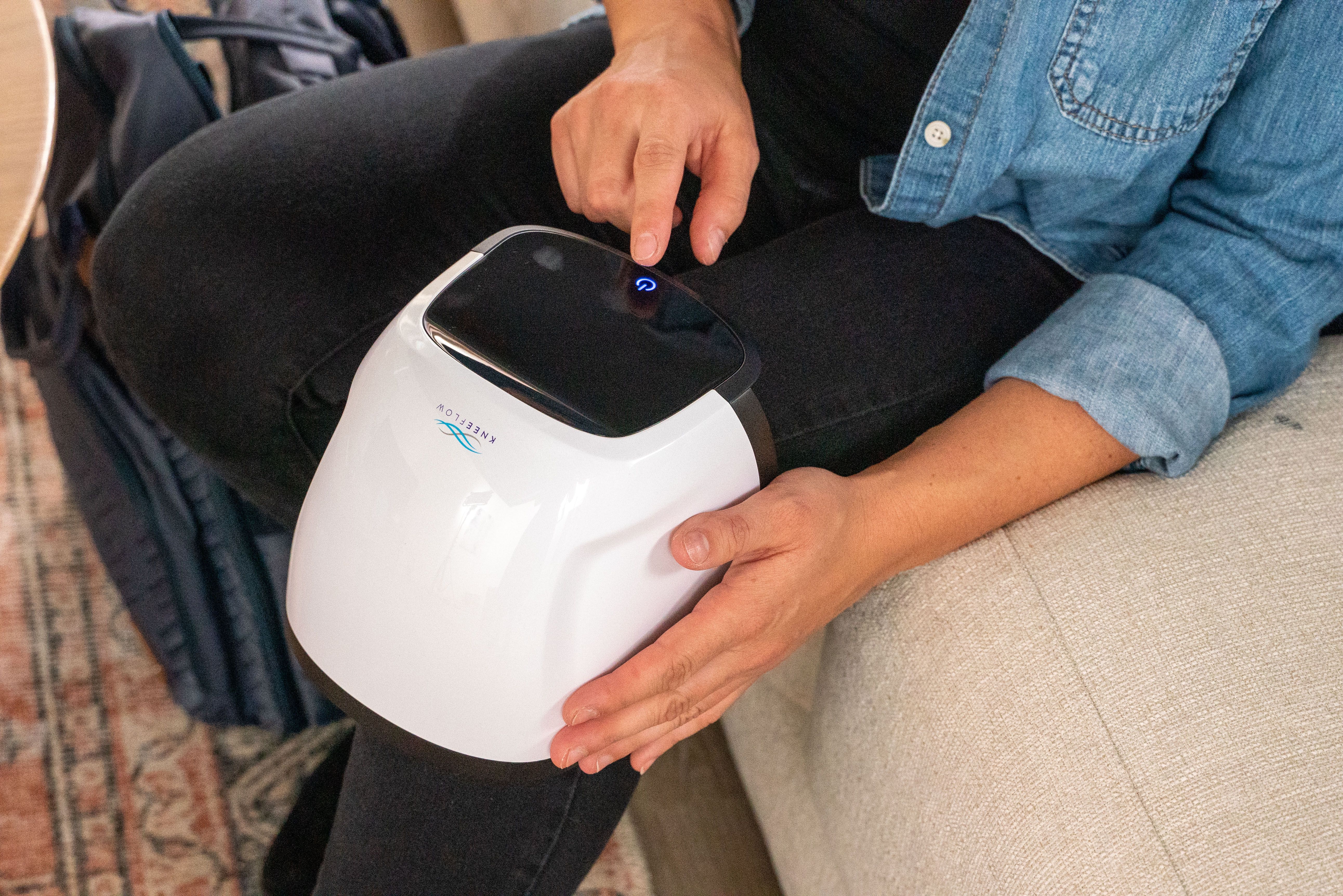 A person presses a blue-lit button on a white Eneflow device sitting on their lap.