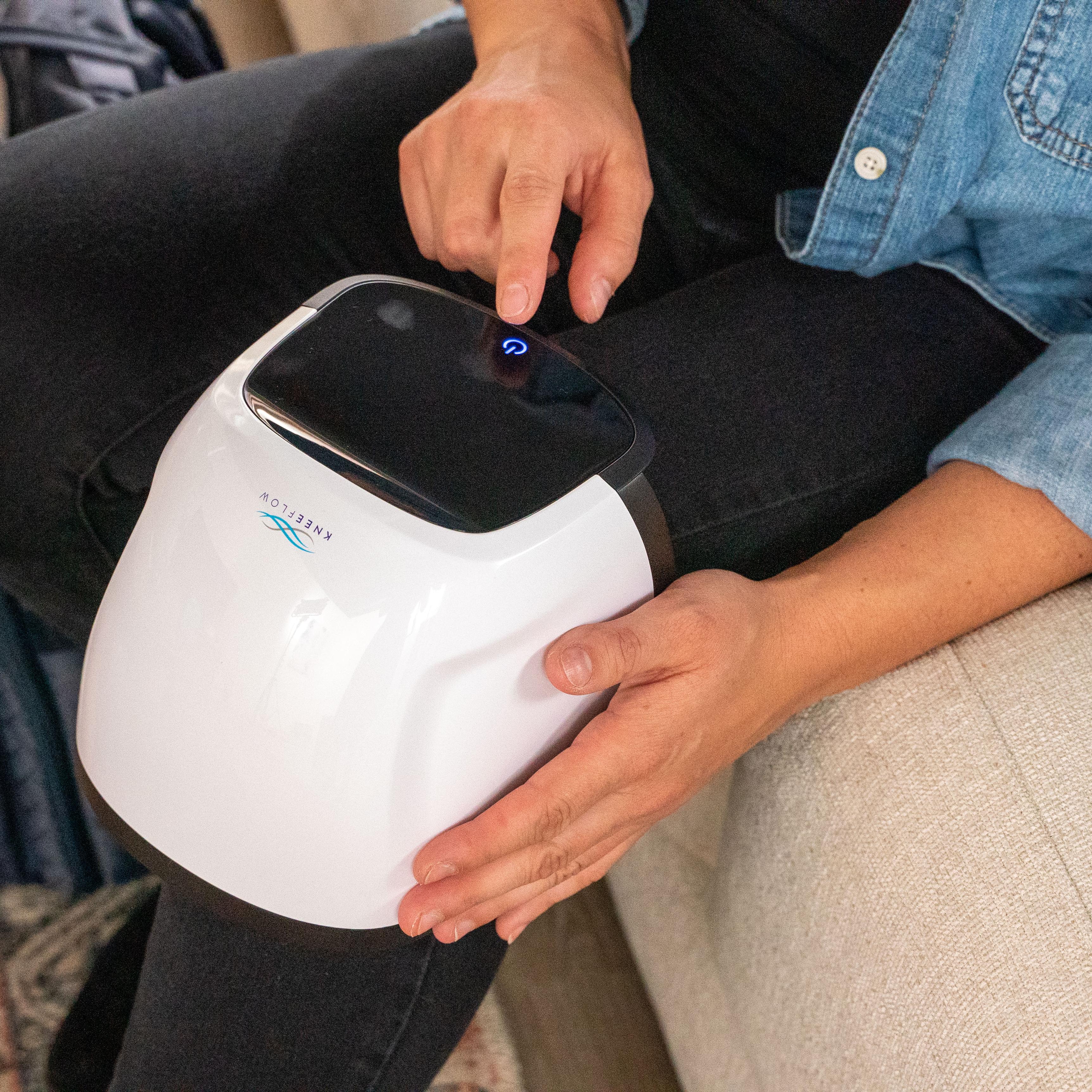 A person presses a blue-lit button on a white Eneflow device sitting on their lap.