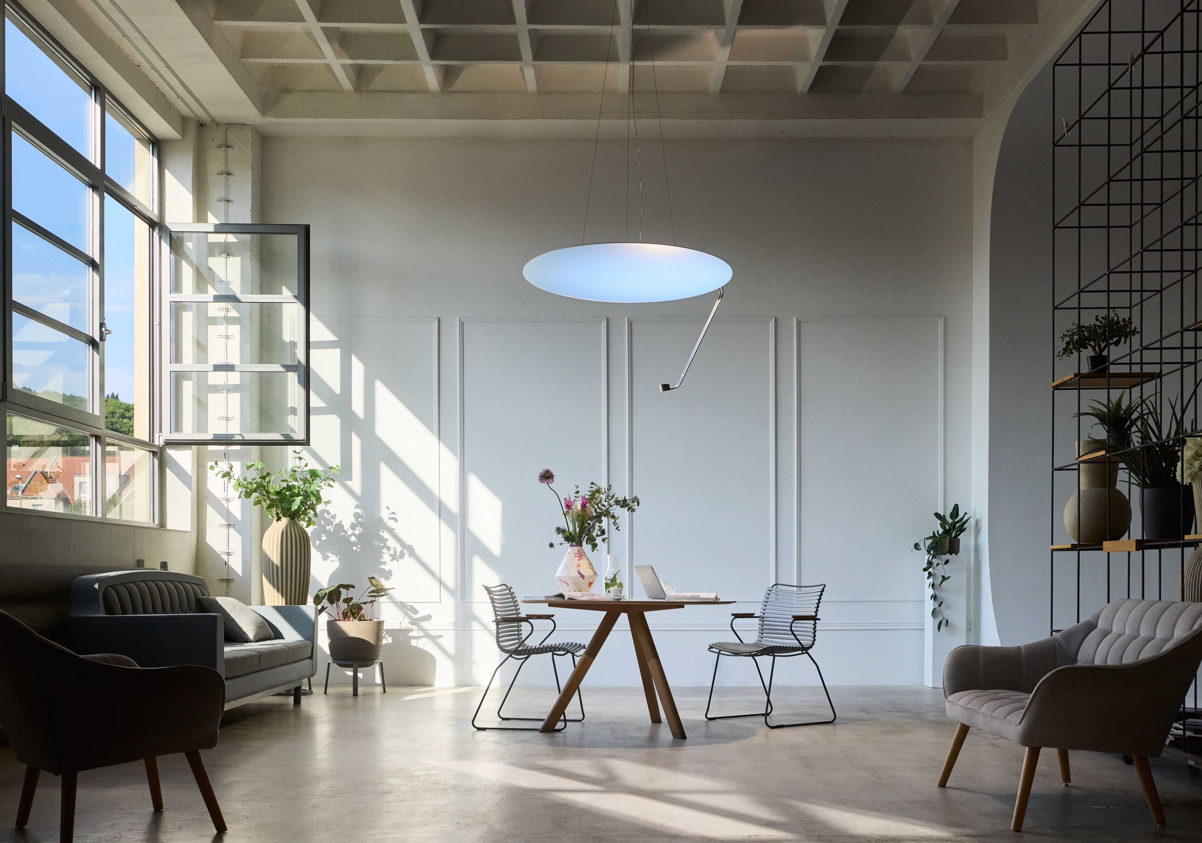 A bright, modern loft space with a wooden table, minimalist chairs, a large circular pendant light, and sunlit windows.
