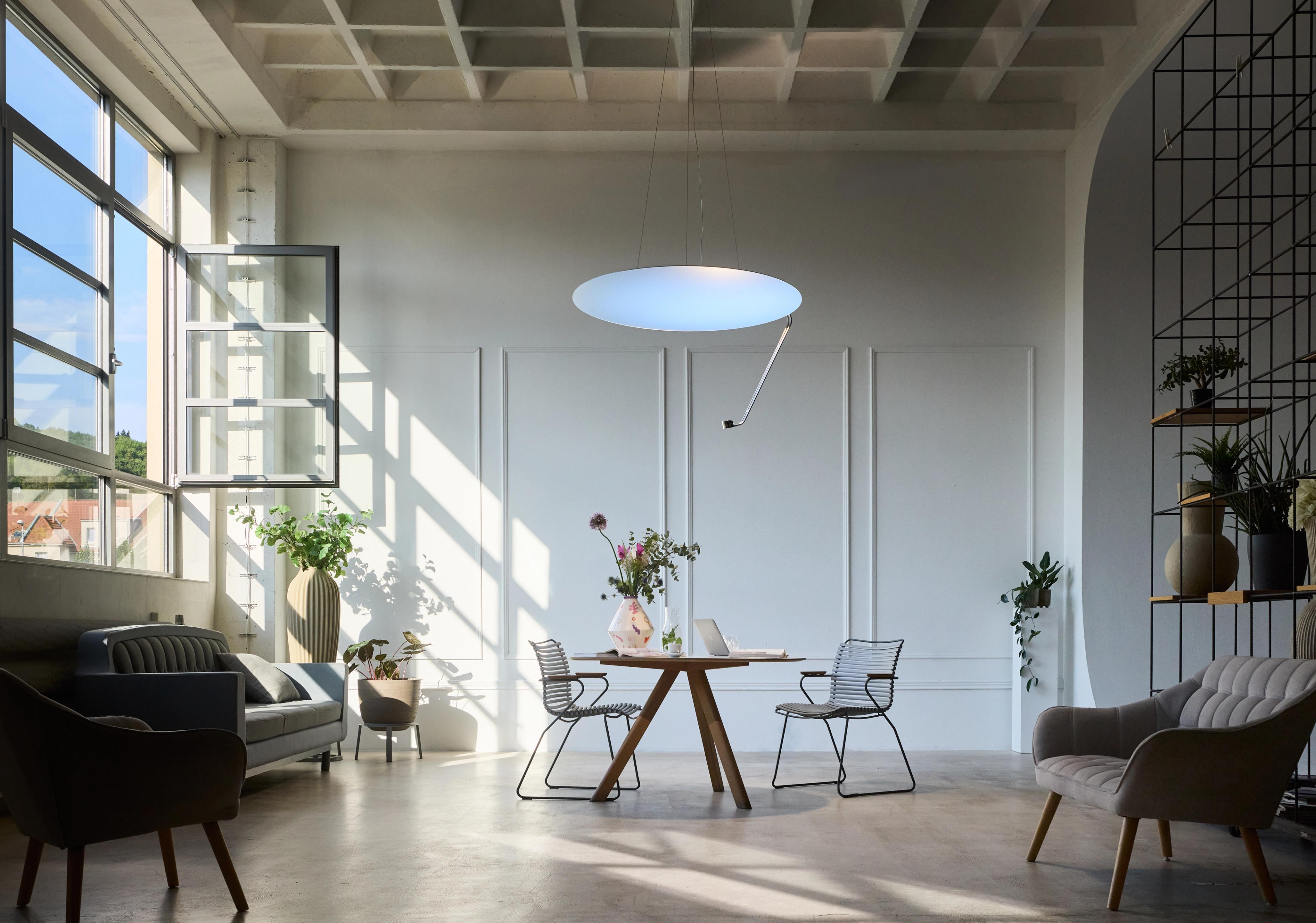 A bright, modern loft space with a wooden table, minimalist chairs, a large circular pendant light, and sunlit windows.