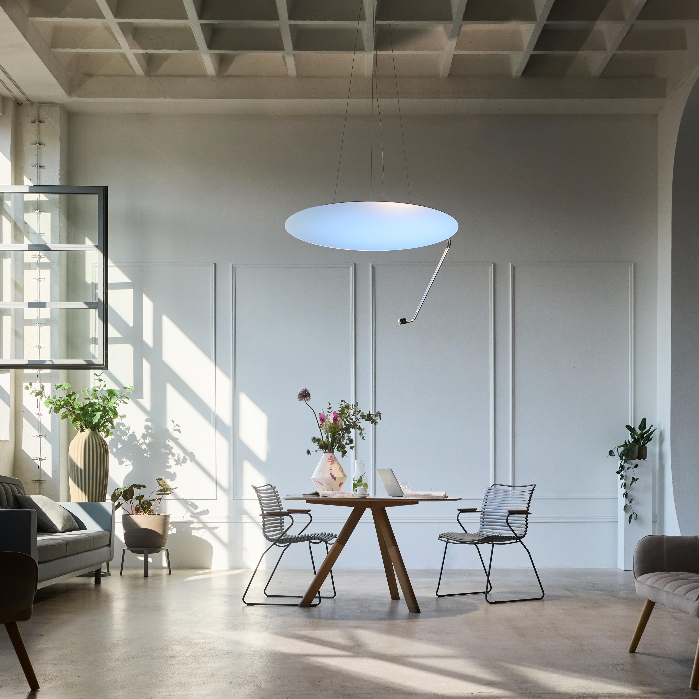 A bright, modern loft space with a wooden table, minimalist chairs, a large circular pendant light, and sunlit windows.