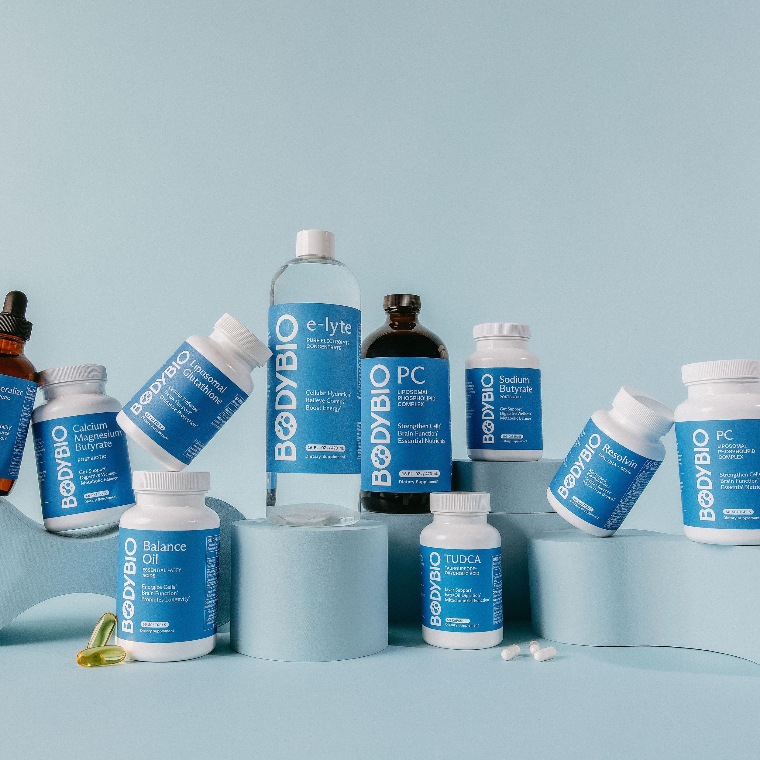 A collection of BodyBio supplement bottles and vials arranged on light blue risers against a light blue background.