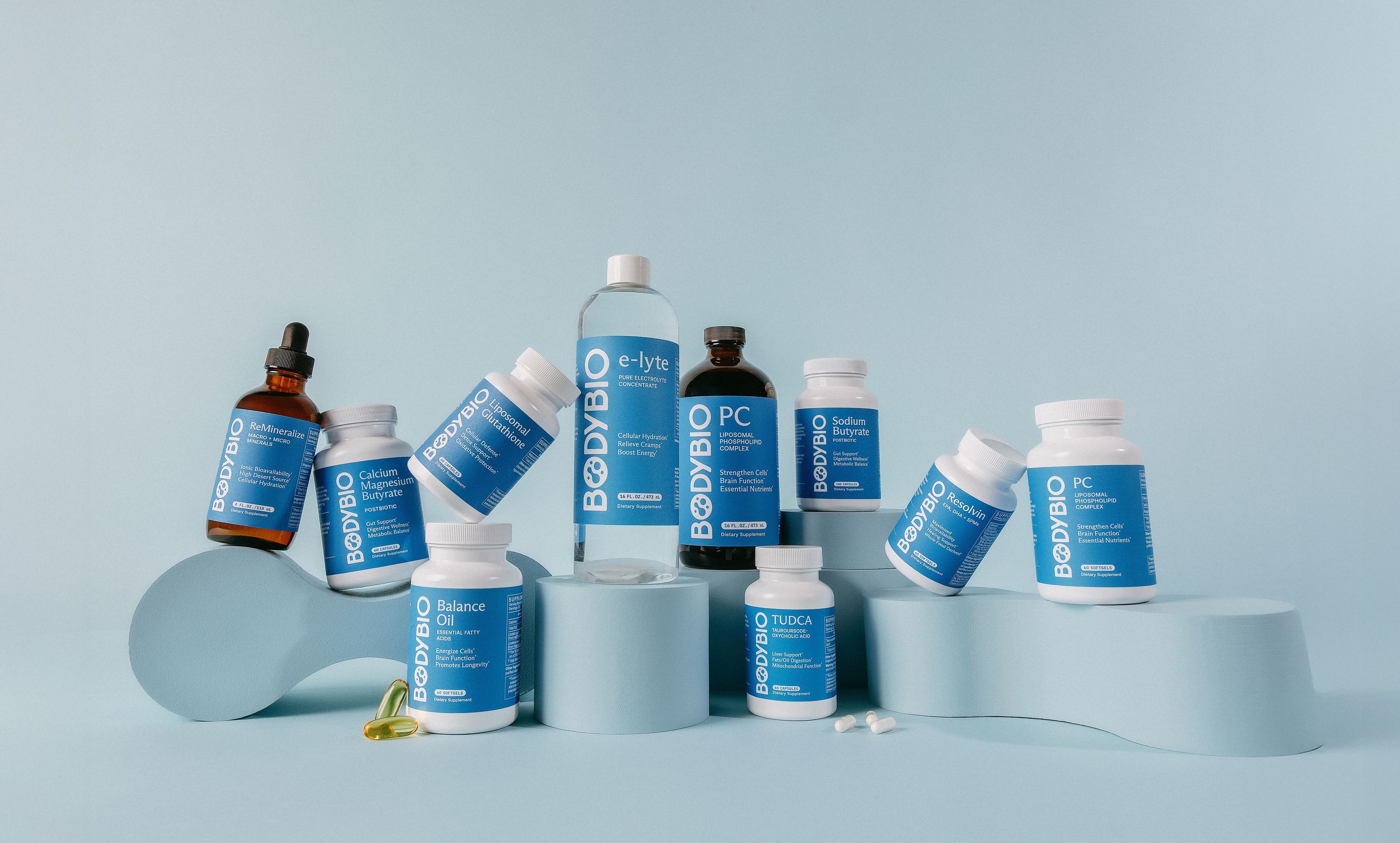 A collection of BodyBio supplement bottles and vials arranged on light blue risers against a light blue background.