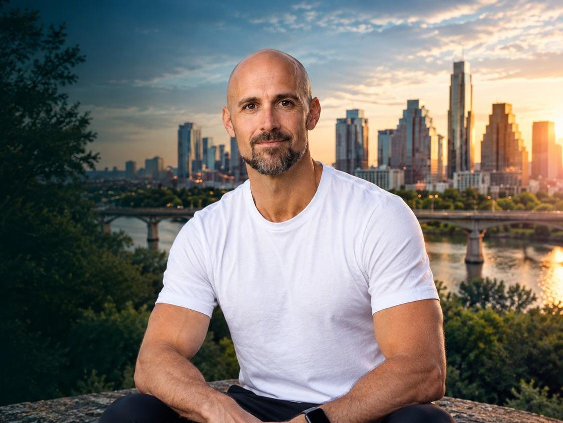 A bald man with a beard in a white t-shirt sits smiling, with a city skyline and river at sunset behind him.
