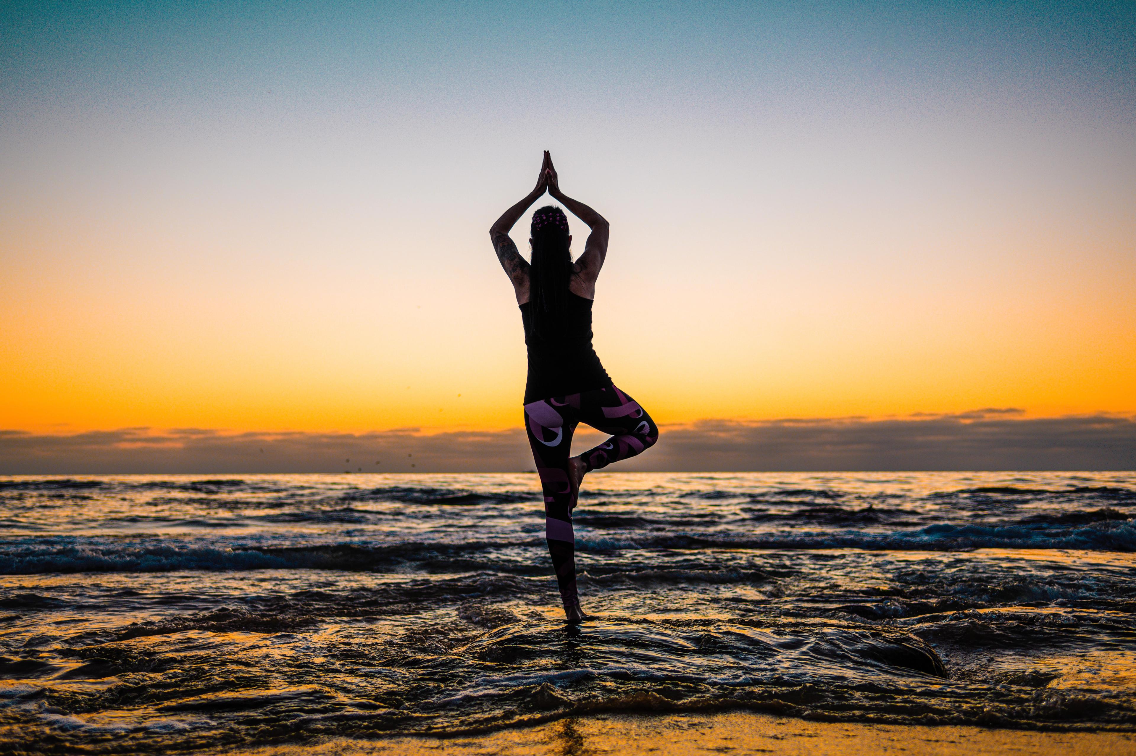 A person in tree pose yoga stands in the ocean at sunset.
