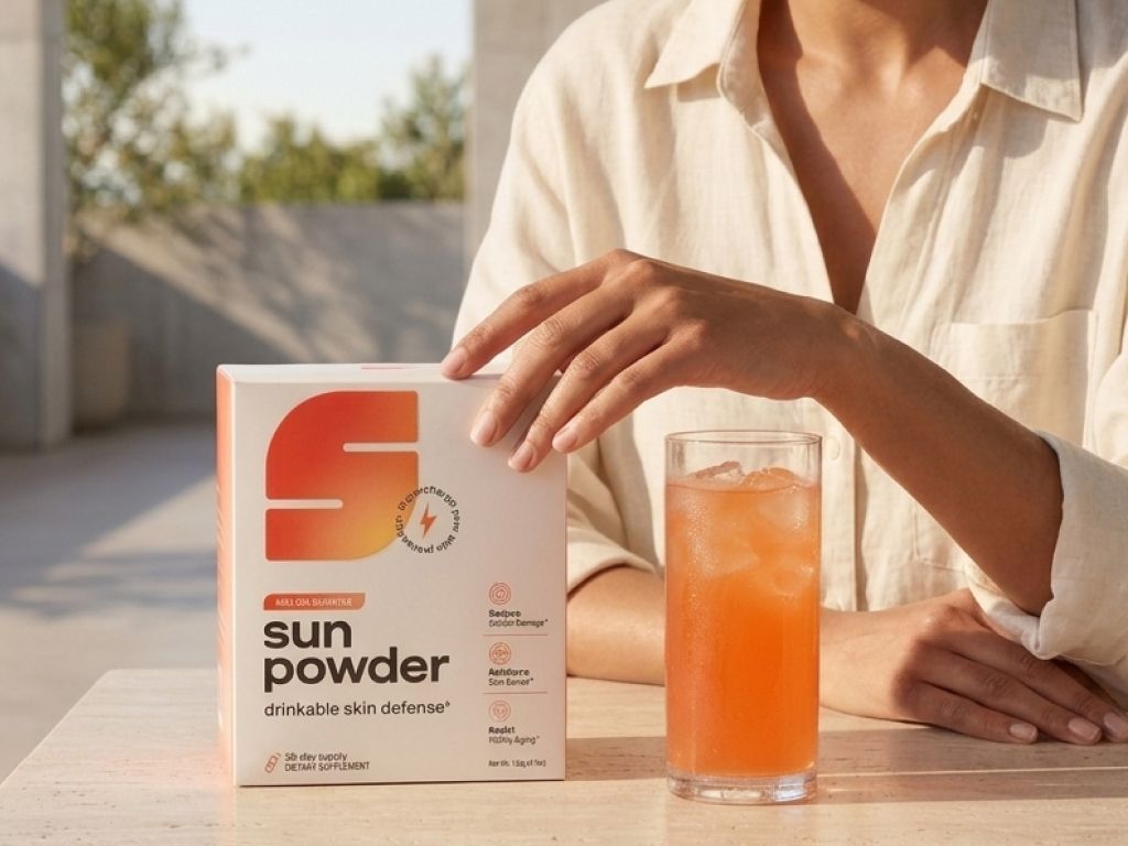 A box of "Sun Powder drinkable skin defense" next to a glass of orange iced drink, with a person's hands.