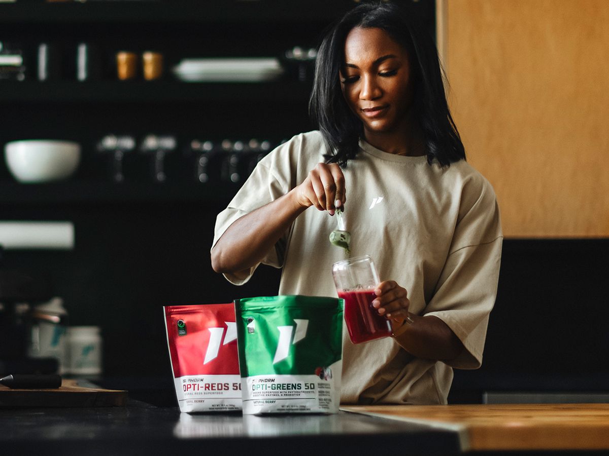 Woman adds green supplement powder to a drink, with 'OPTI-REDS' and 'OPTI-GREENS' bags on the counter.