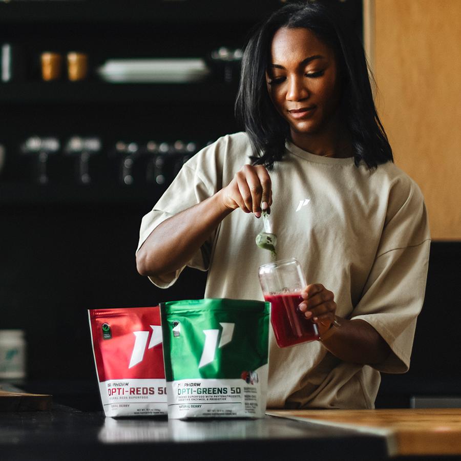 Woman adds green supplement powder to a drink, with 'OPTI-REDS' and 'OPTI-GREENS' bags on the counter.