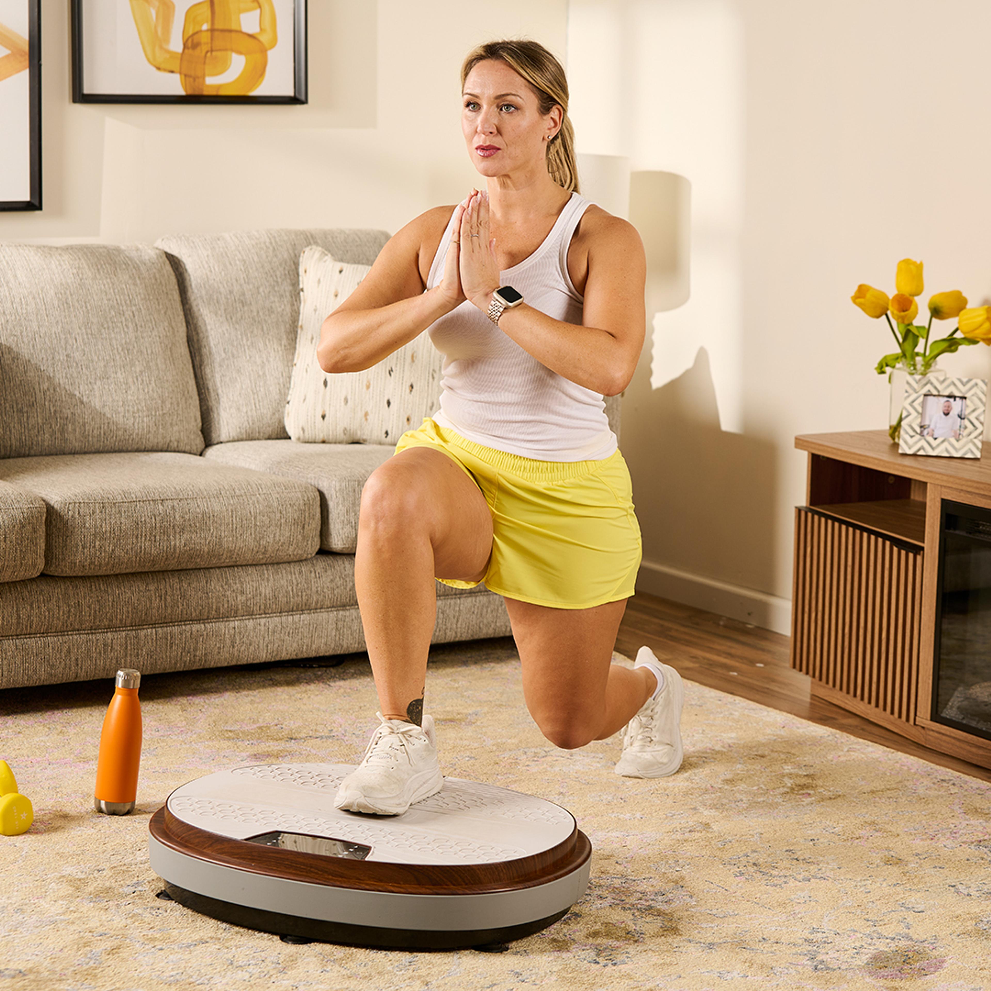 A woman lunges on a vibration platform in a living room.