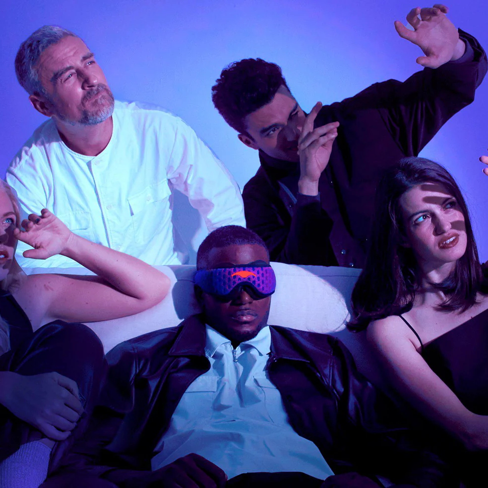 Five people on a couch; one man wears a futuristic eye mask, while others react to a dramatic purple light.