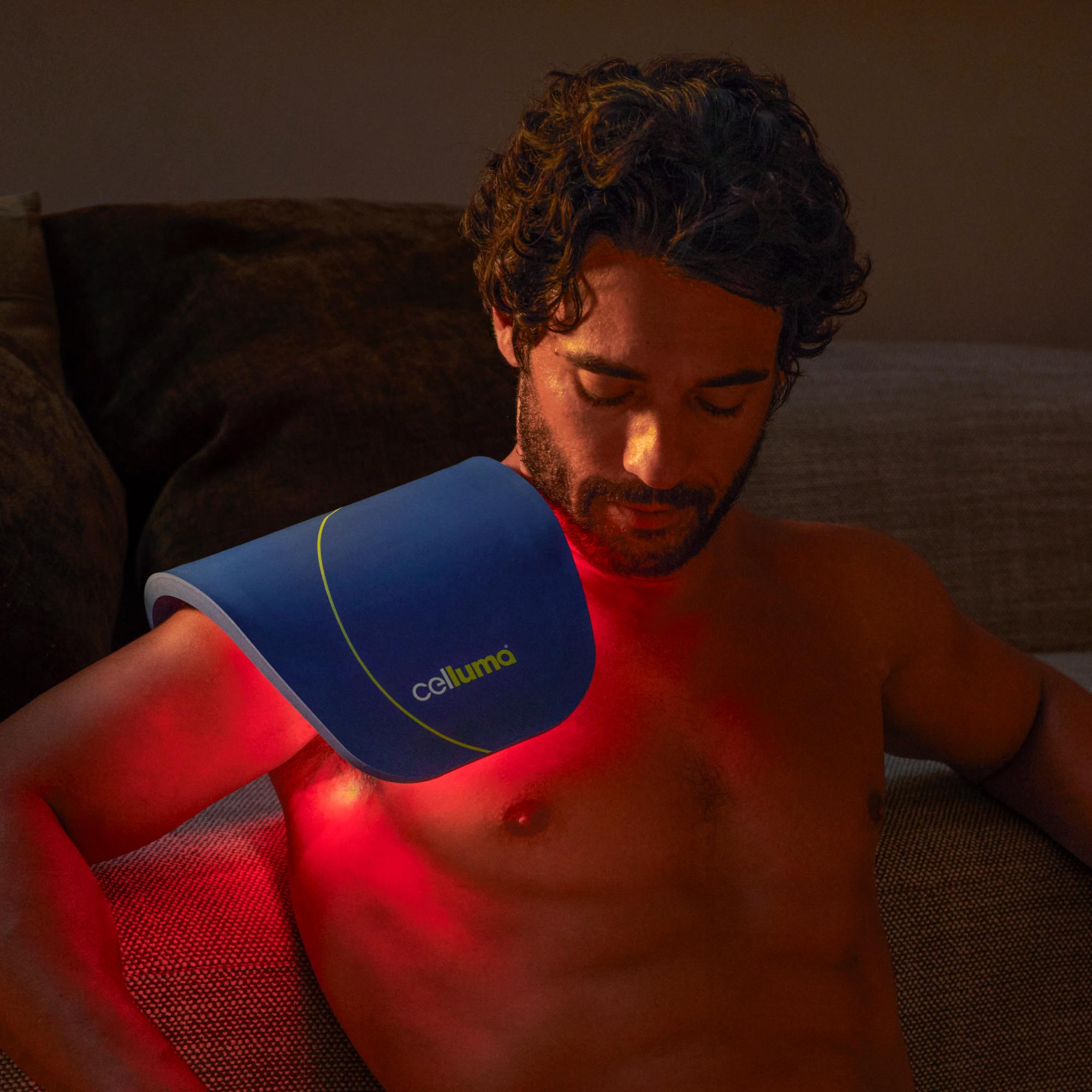 A man applies a blue Celluma red light therapy device to his shoulder, illuminating his skin with red light.