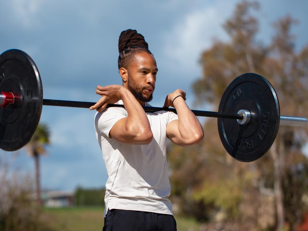 Man with dreadlocks holding a barbell in a front rack position outdoors.