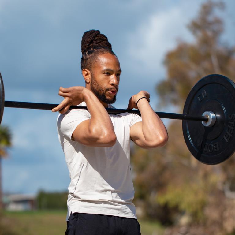 Man with dreadlocks holding a barbell in a front rack position outdoors.