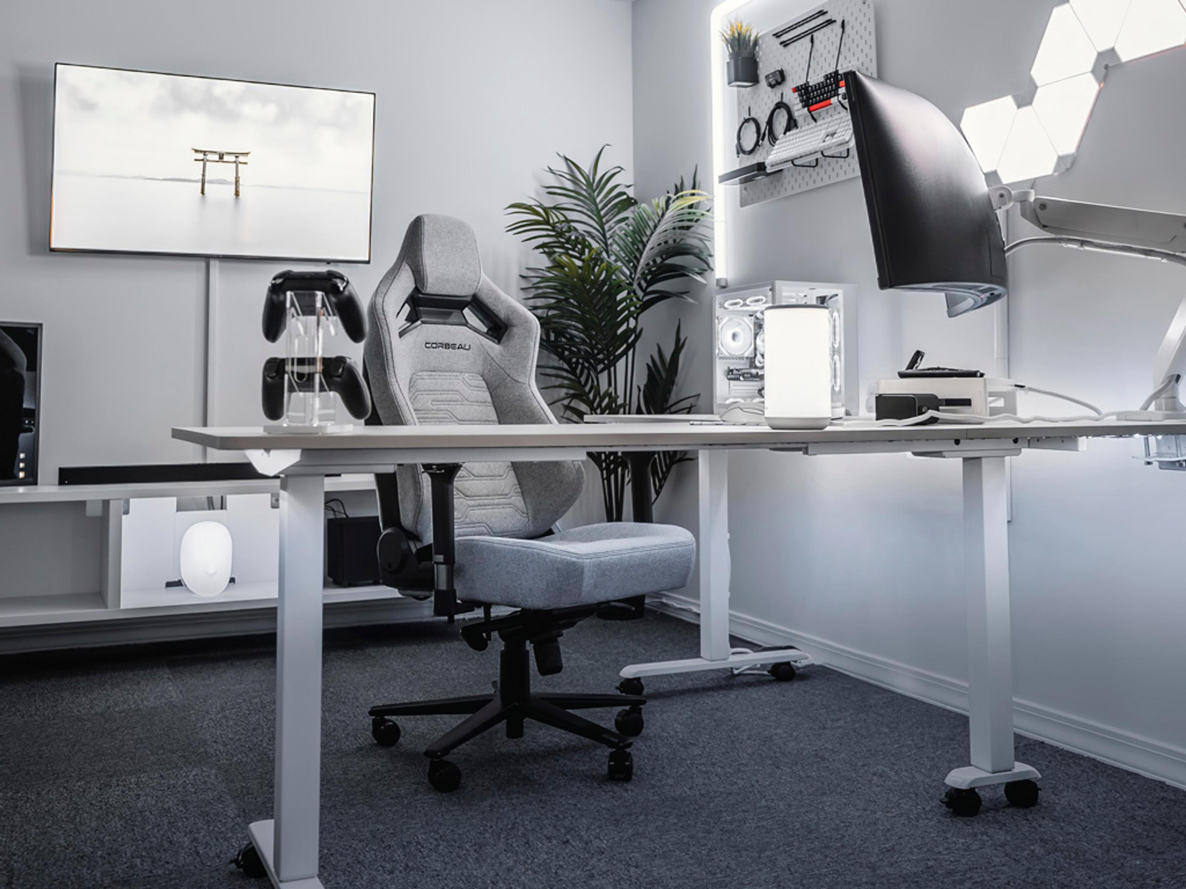 A modern, monochromatic white and grey gaming setup with a standing desk, gaming chair, large TV, desktop PC, and geometric wall lights.