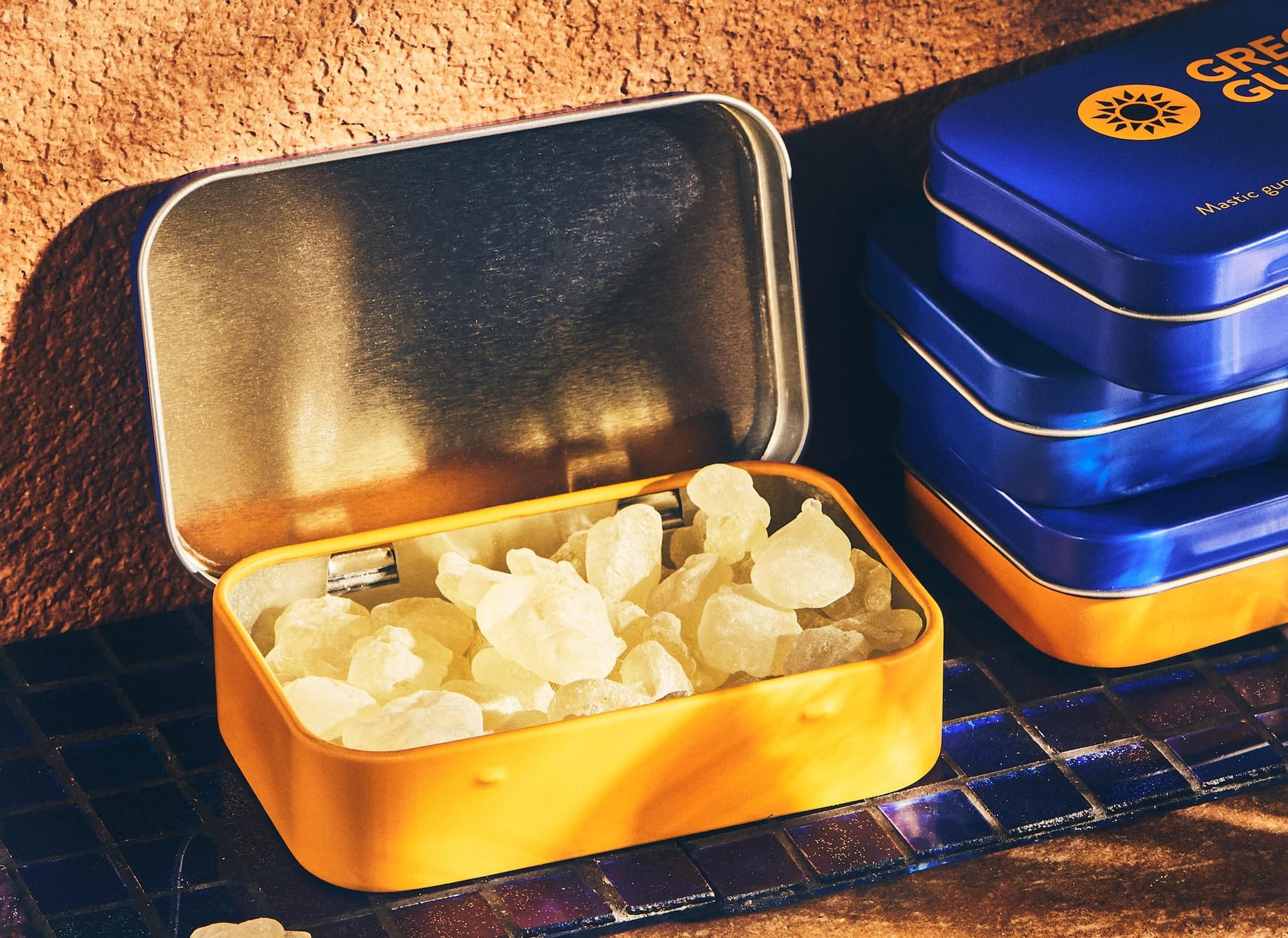 An open orange tin filled with pale yellow mastic gum pieces, next to a stack of blue and orange branded tins.