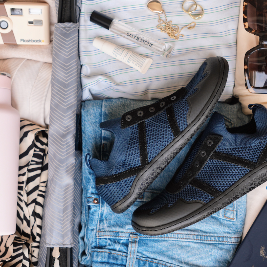 An open suitcase packed with blue mesh sneakers, folded jeans, a camera, sunglasses, and toiletries.