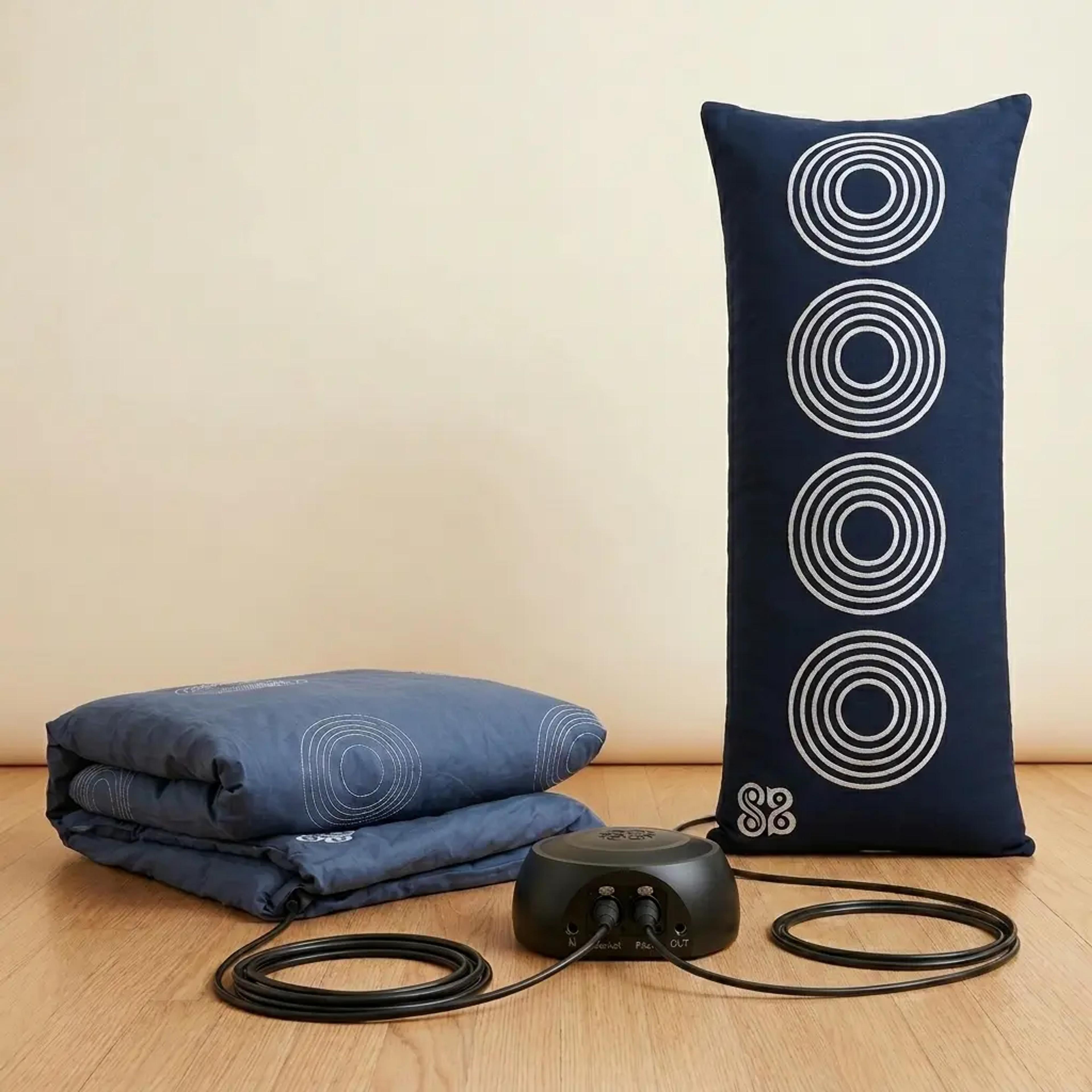 A folded blue mattress pad and an upright dark blue body pillow with white concentric circles are connected to a black control unit by cables on a wooden floor.