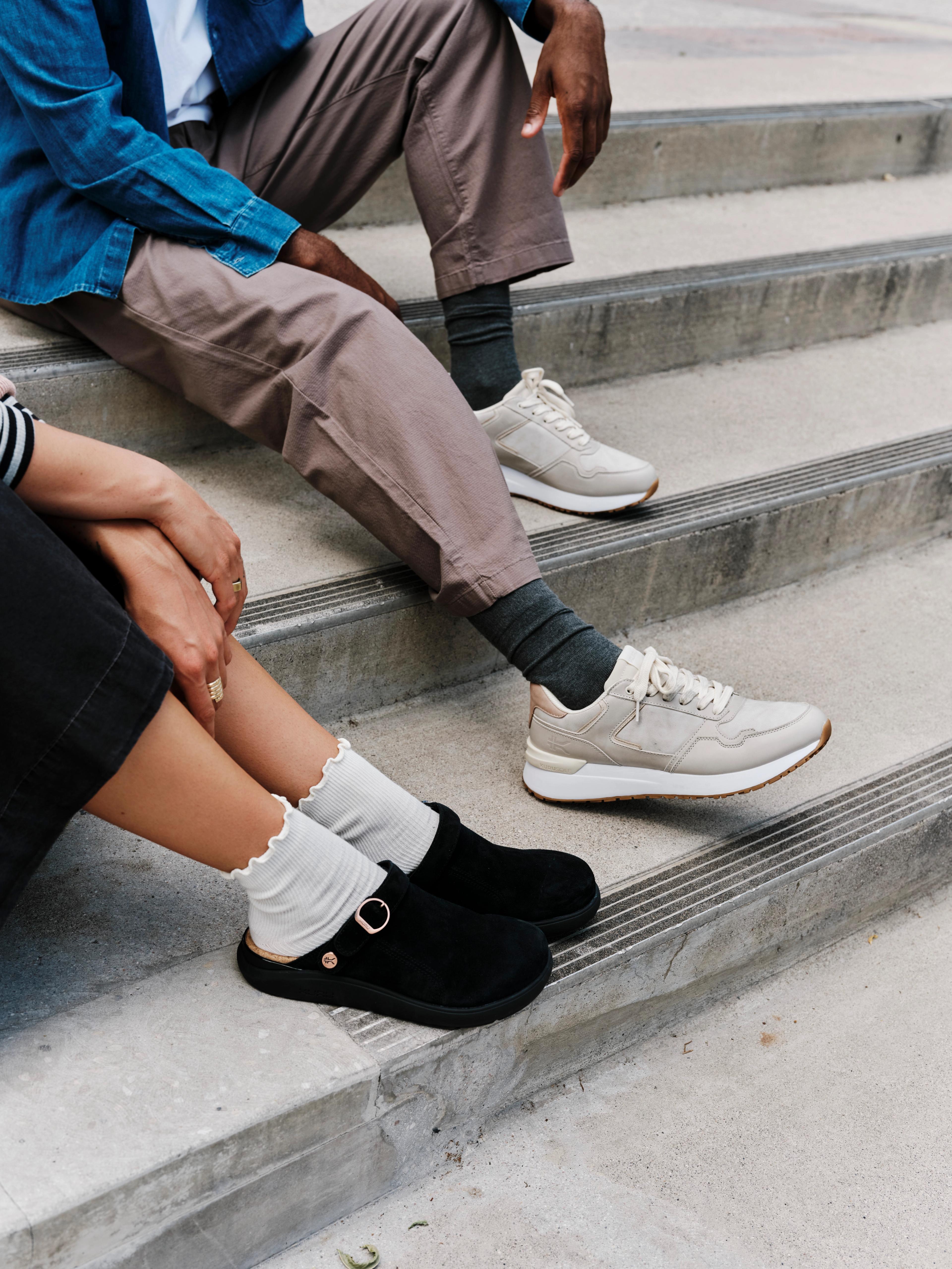 Two people sit on concrete stairs, one in beige sneakers and the other in black clogs with white ruffled socks.