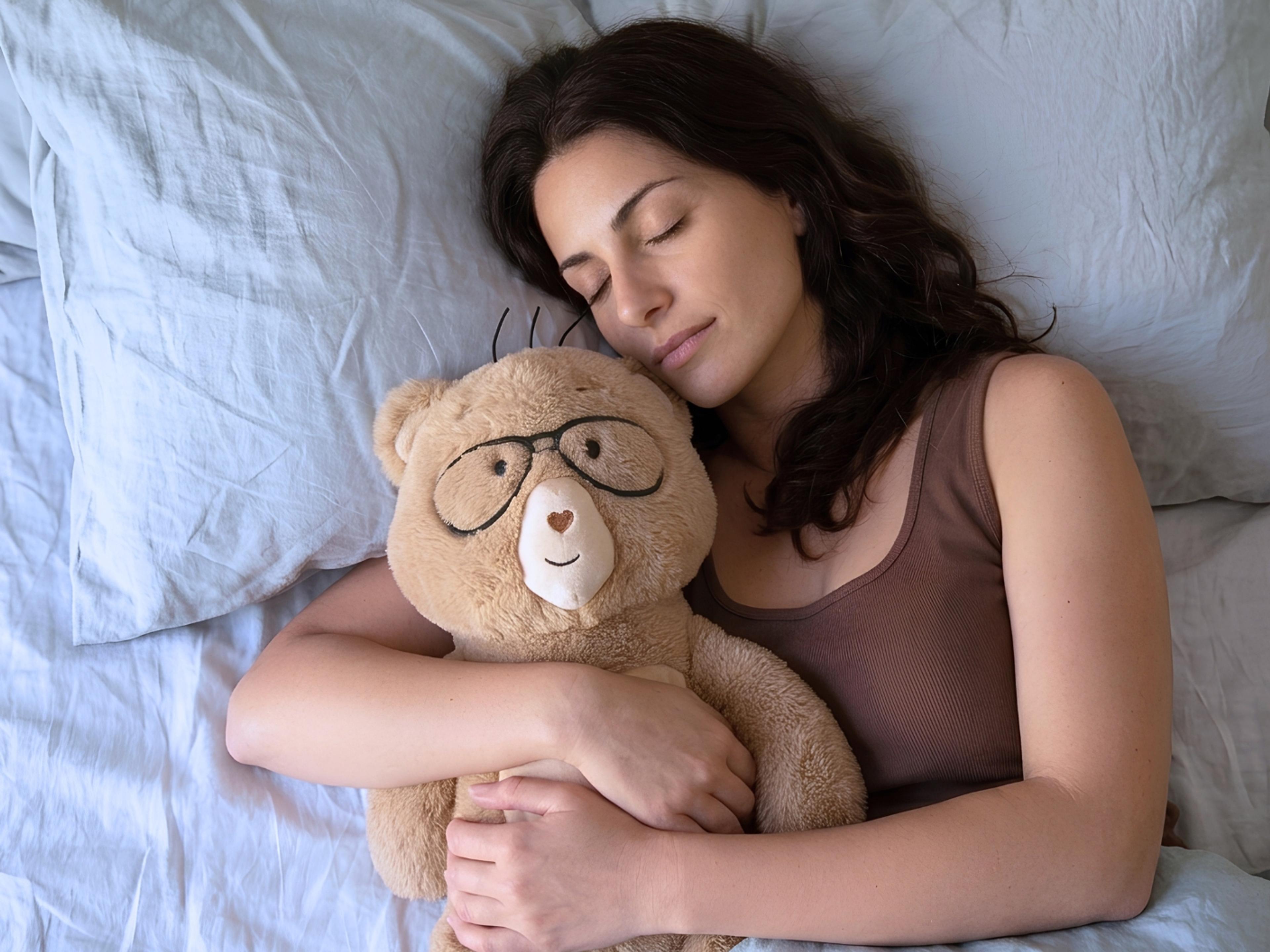A woman sleeping soundly in bed, hugging a teddy bear with glasses.