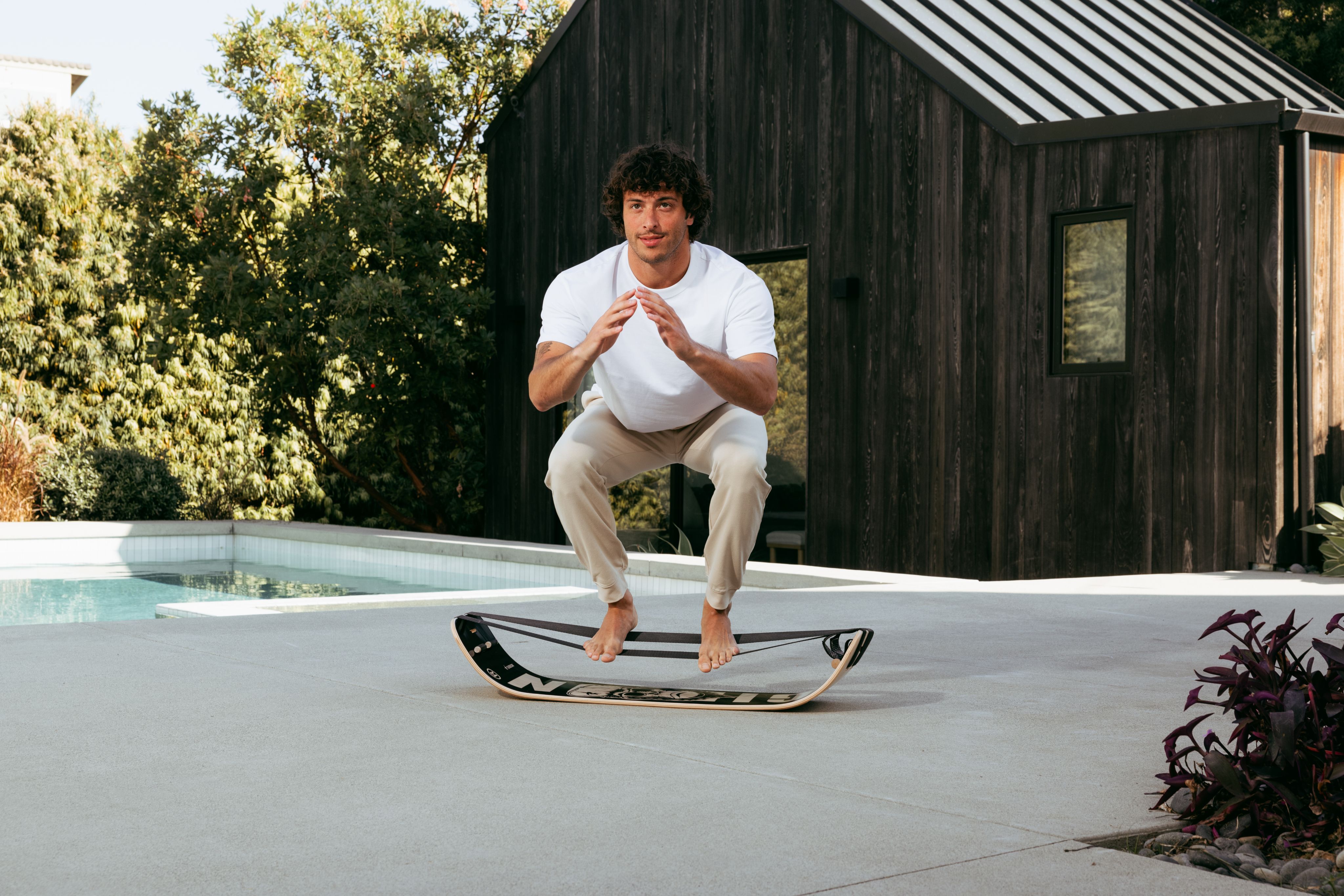 Man in a dynamic squat above a balance board outdoors.