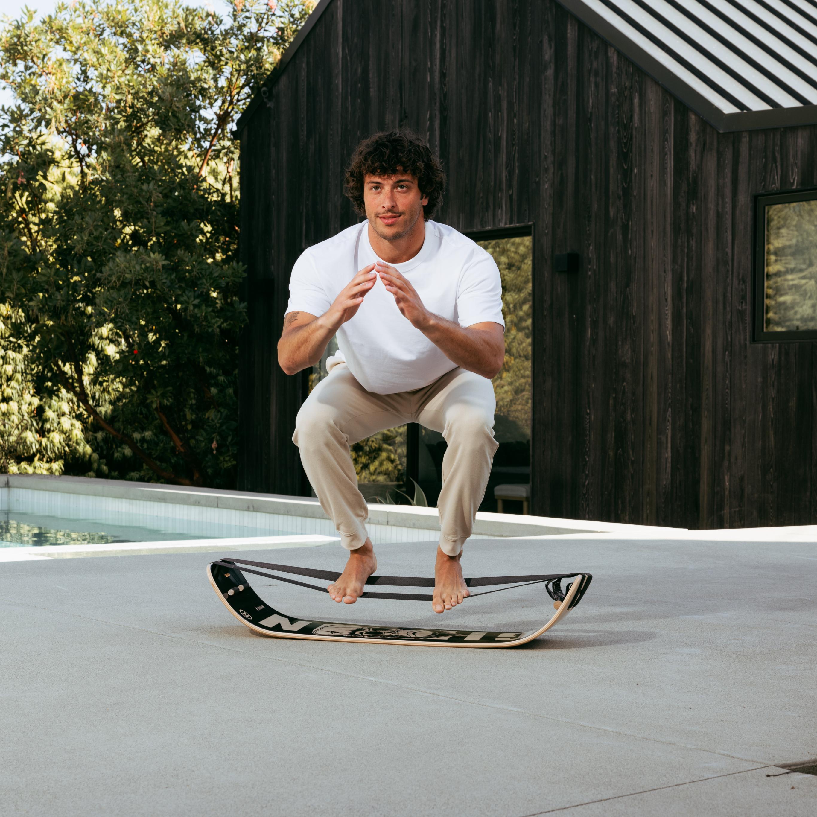 Man in a dynamic squat above a balance board outdoors.