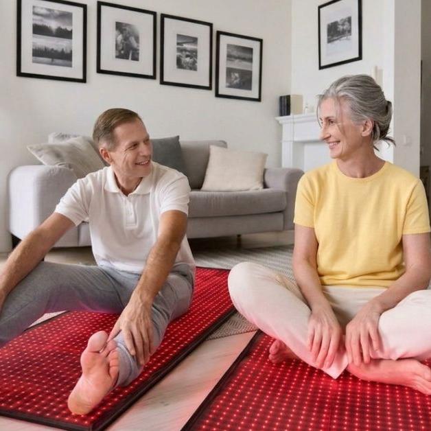 An older couple smiles while relaxing on red light therapy mats in their living room.