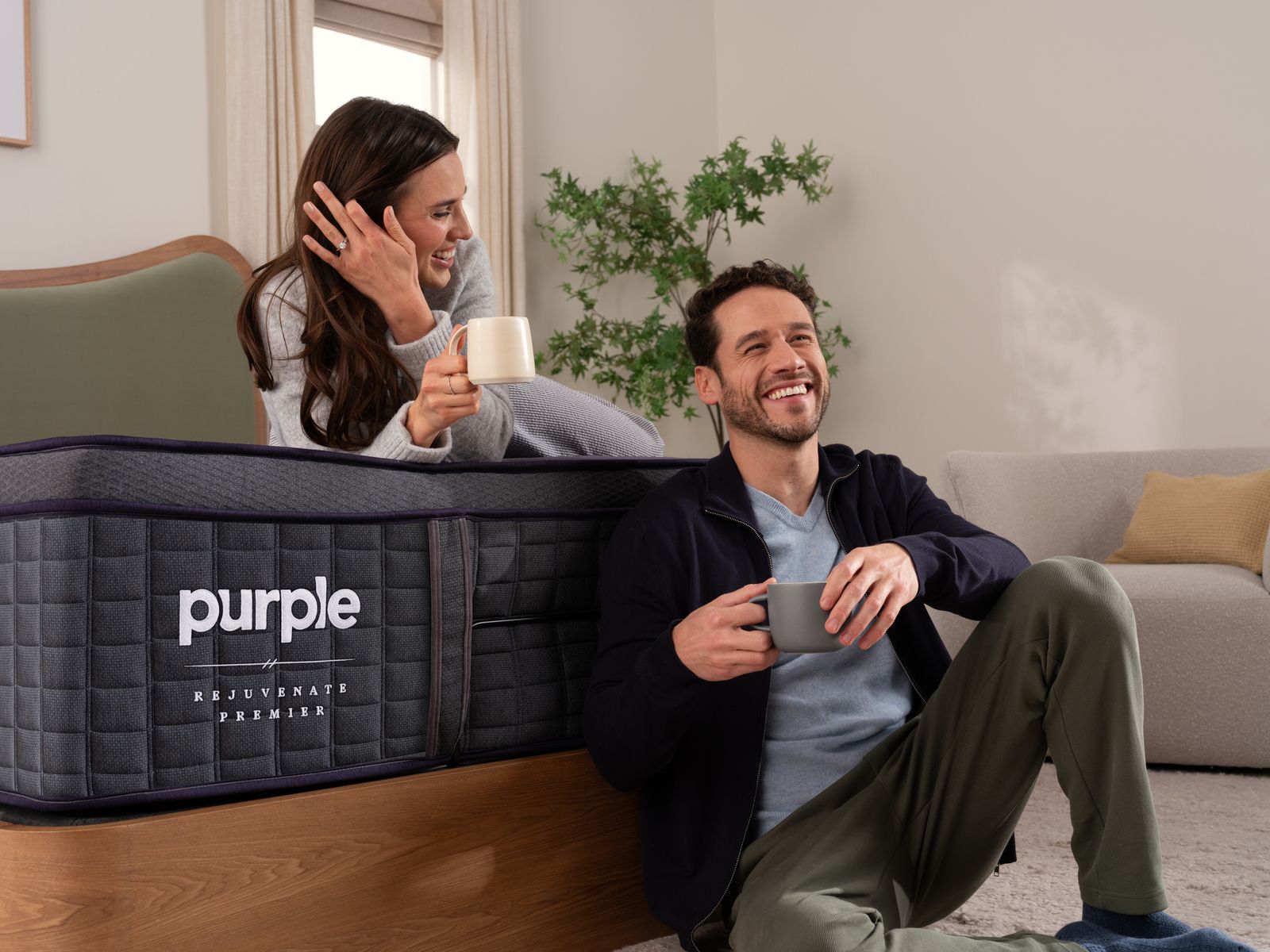 Couple laughing and holding mugs while resting on Purple mattress