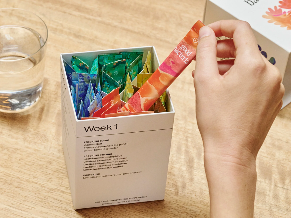 A hand removes a colorful probiotic supplement packet from a box labeled "Week 1", with a glass of water nearby.