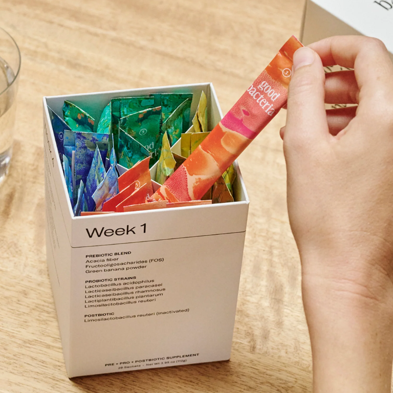 A hand removes a colorful probiotic supplement packet from a box labeled "Week 1", with a glass of water nearby.