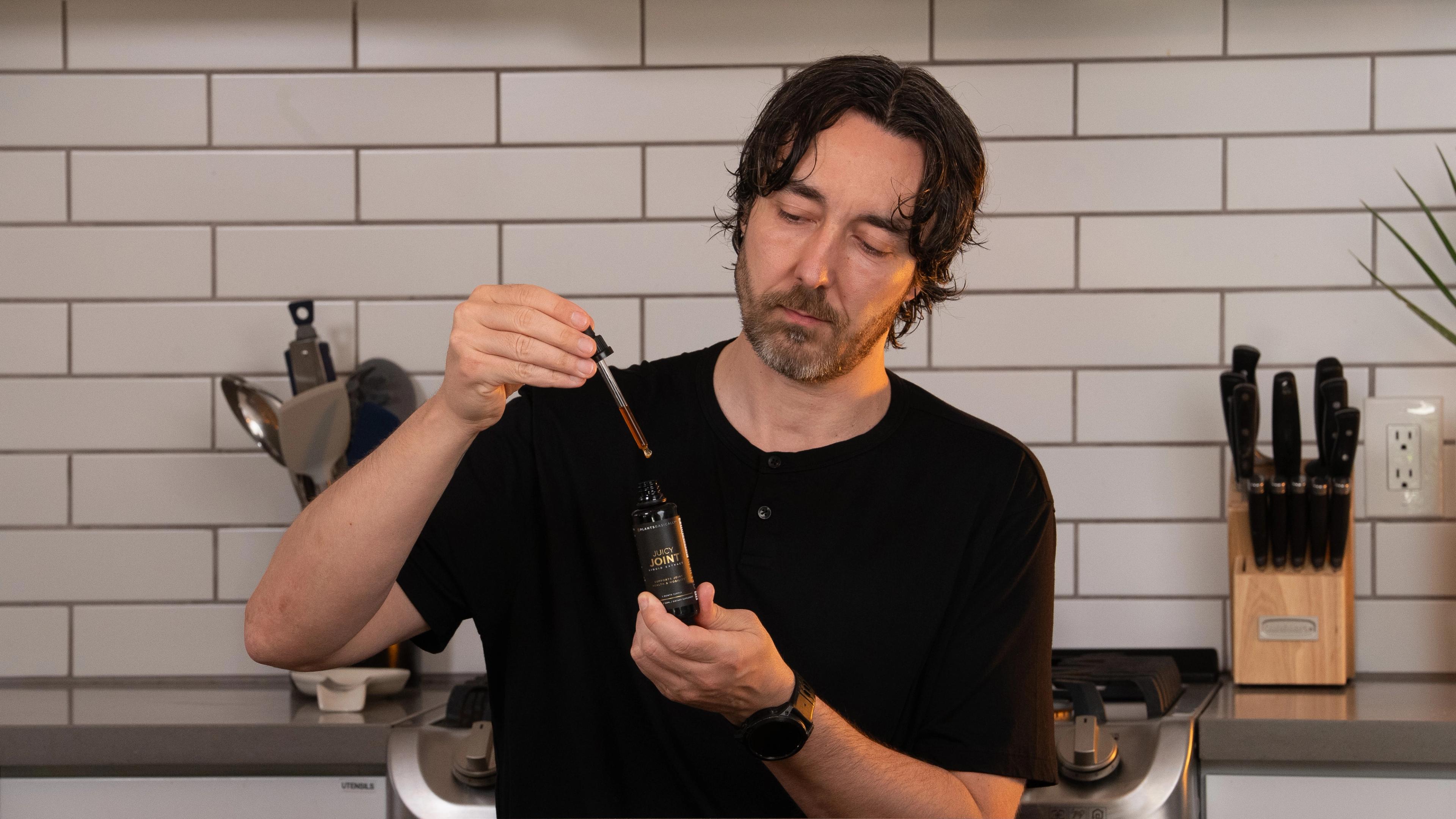 A man holds a dropper bottle, with liquid dripping from the dropper, in a kitchen.