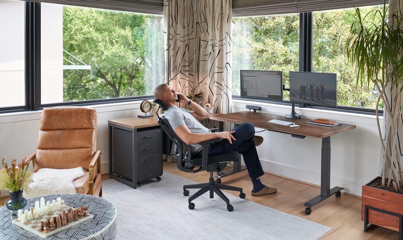 A man talks on a phone while relaxing in an office chair at a standing desk with dual monitors in a modern home office with large windows.