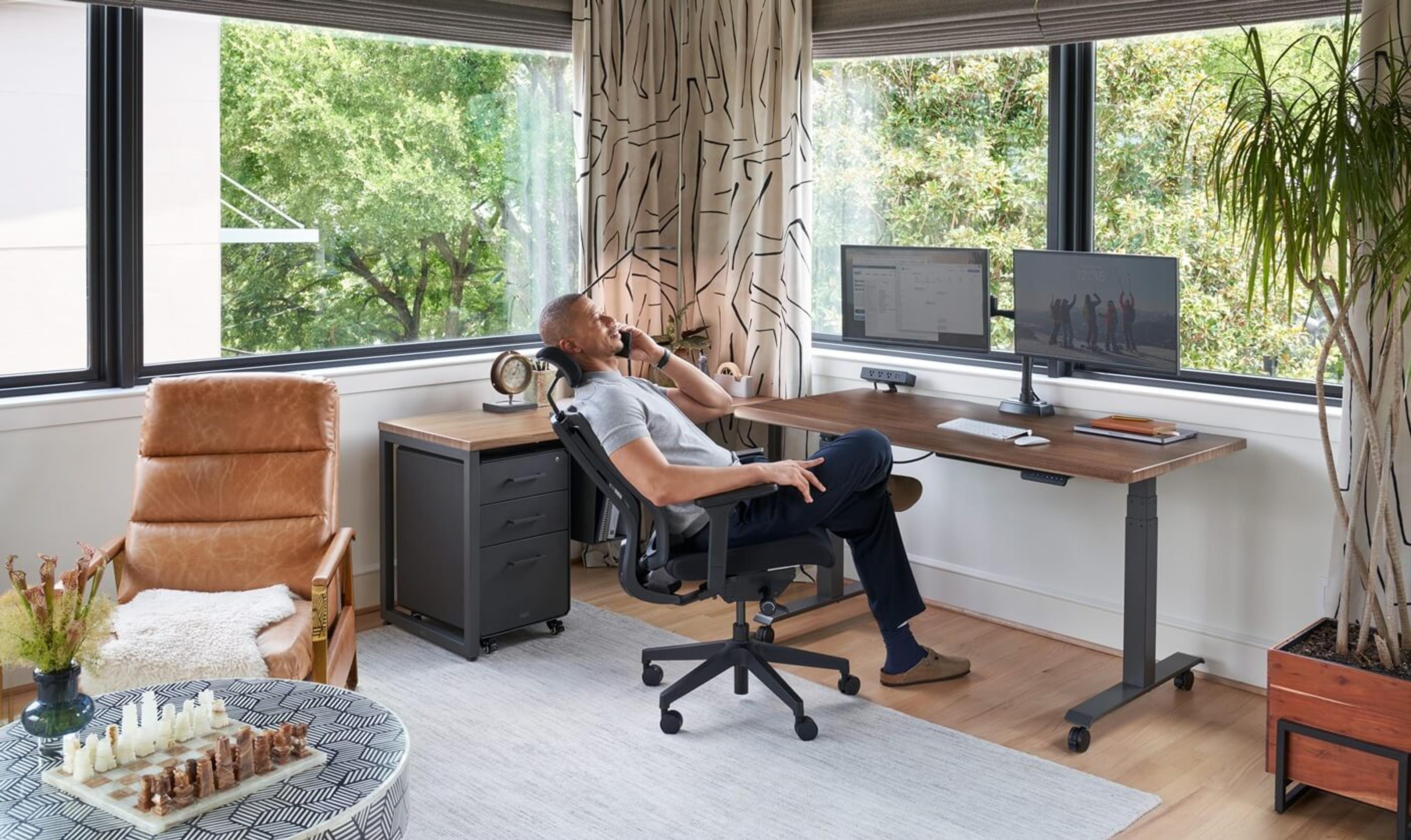 A man talks on a phone while relaxing in an office chair at a standing desk with dual monitors in a modern home office with large windows.