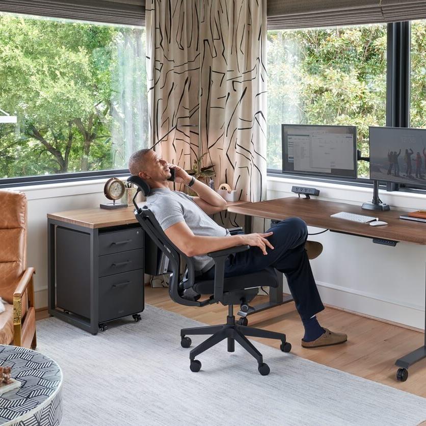 A man talks on a phone while relaxing in an office chair at a standing desk with dual monitors in a modern home office with large windows.