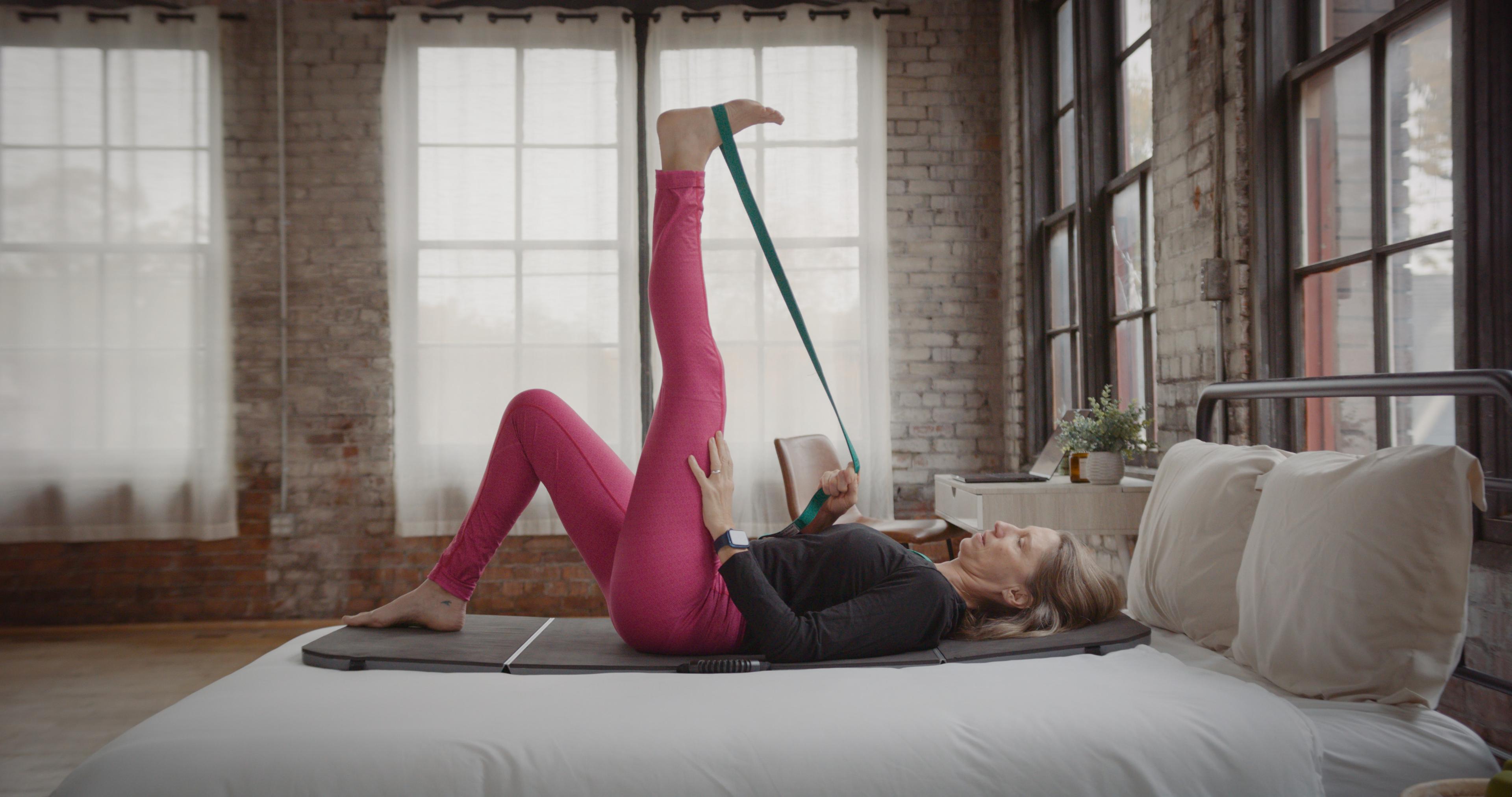 A woman lies on a bed, using a strap to stretch one leg vertically.