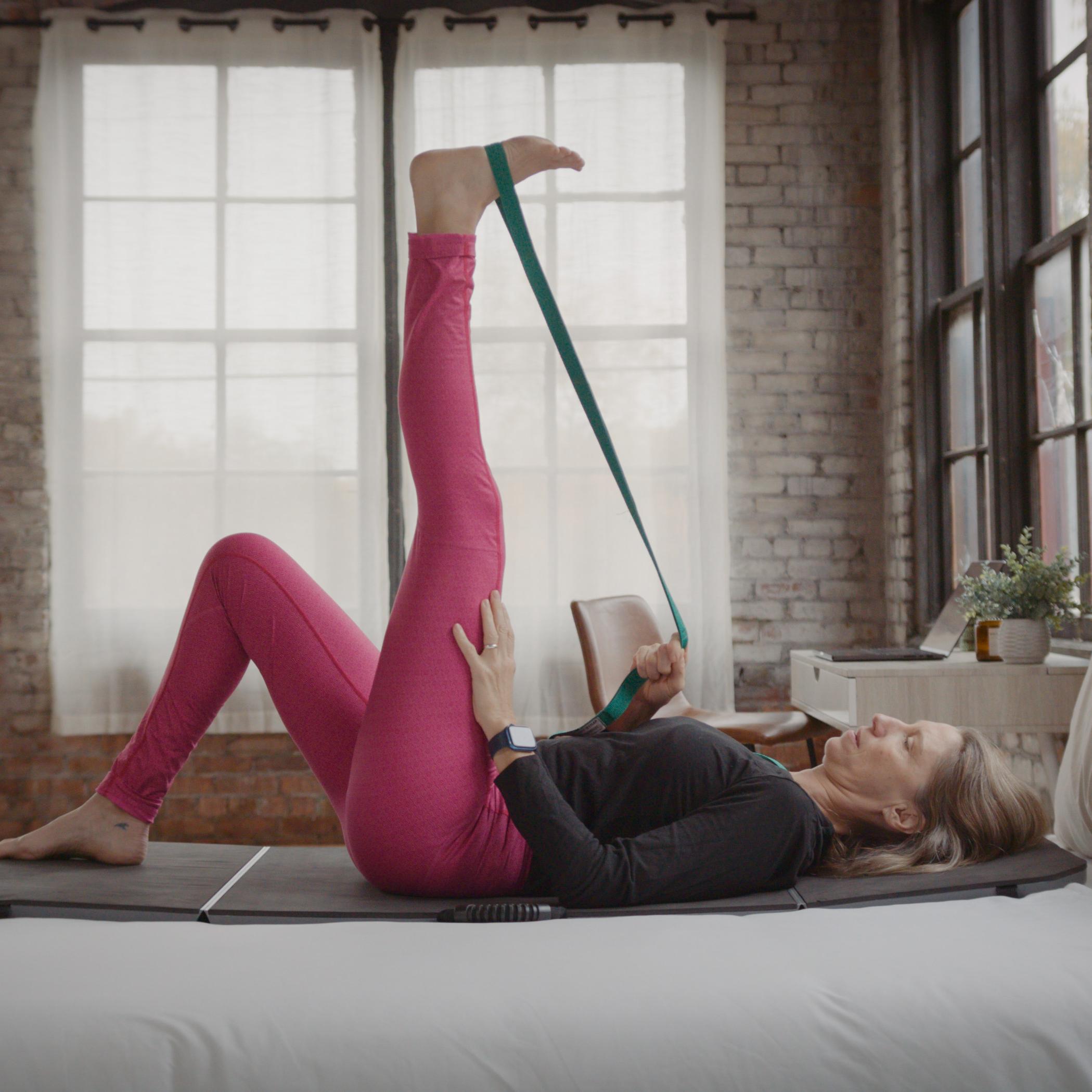 A woman lies on a bed, using a strap to stretch one leg vertically.