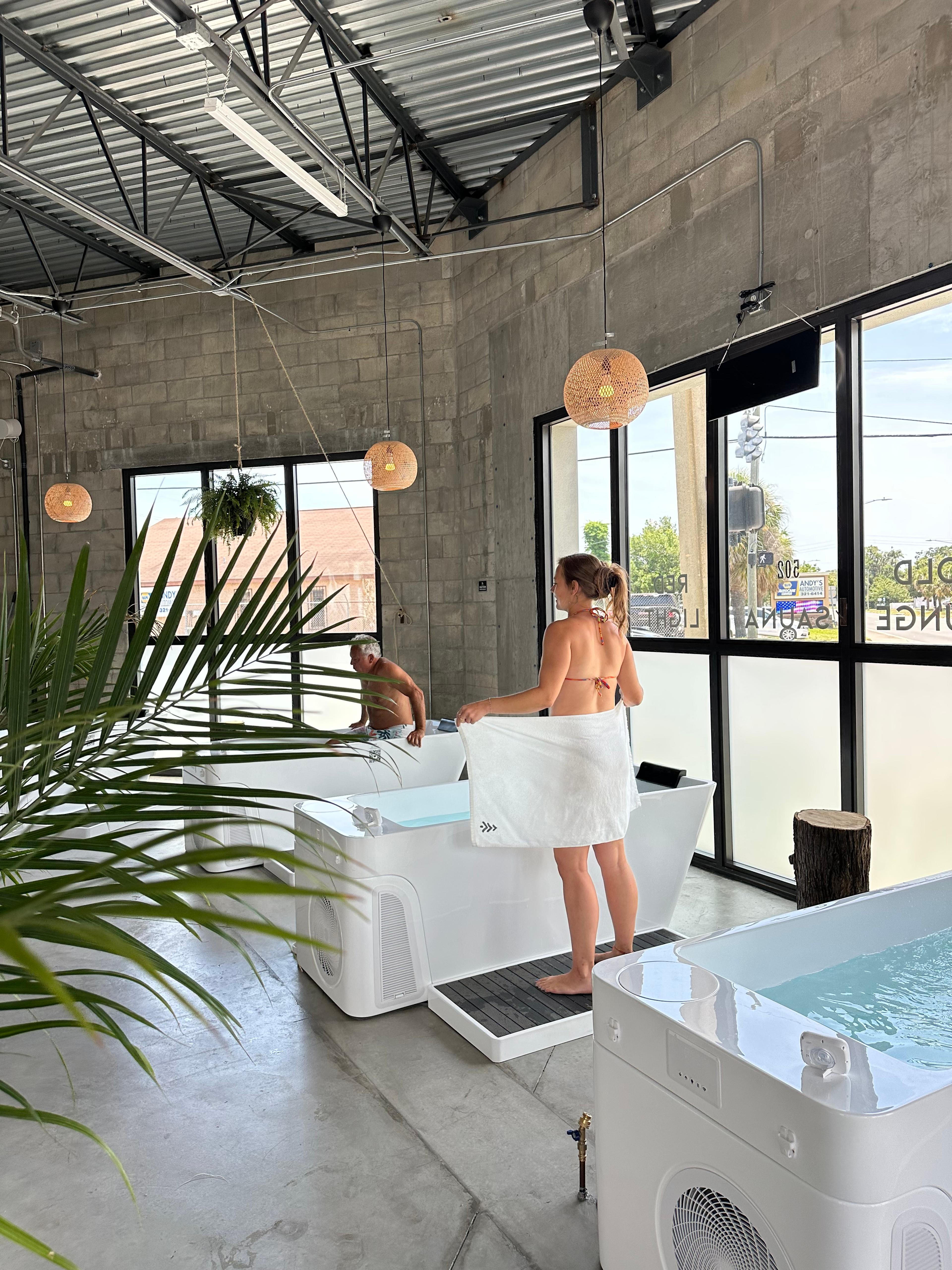 A woman in a bikini and towel stands beside a modern cold plunge tub, with a man in another tub behind her, in a minimalist wellness space.