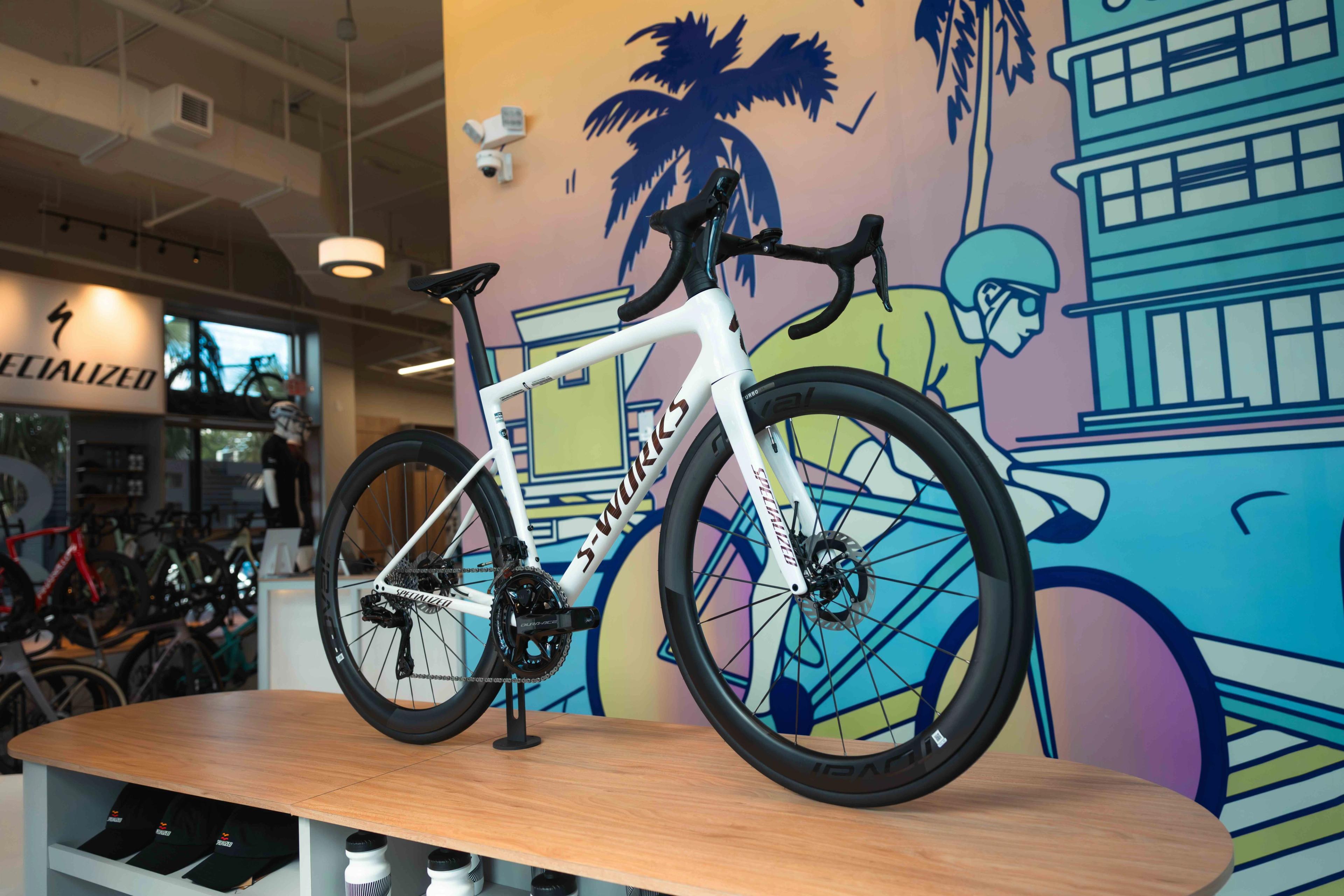 White Specialized S-Works road bike on a wooden display table in a modern bike shop with a colorful mural.
